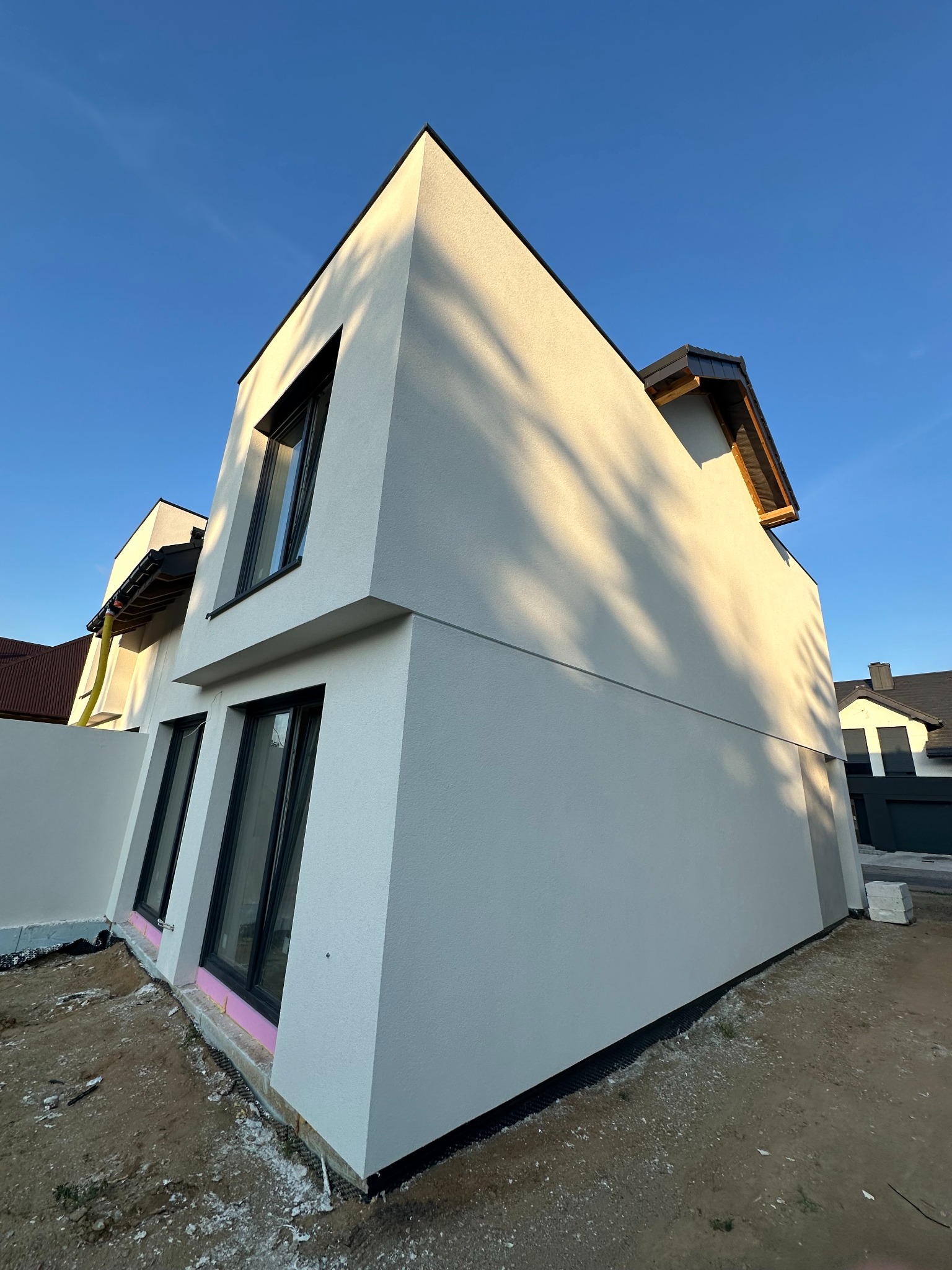 Nowoczesny dom z jasną elewacją w minimalistycznym stylu, czarne ramy okienne, widoczne detale wykończenia, błękitne niebo w tle, cień drzewa na ścianie.