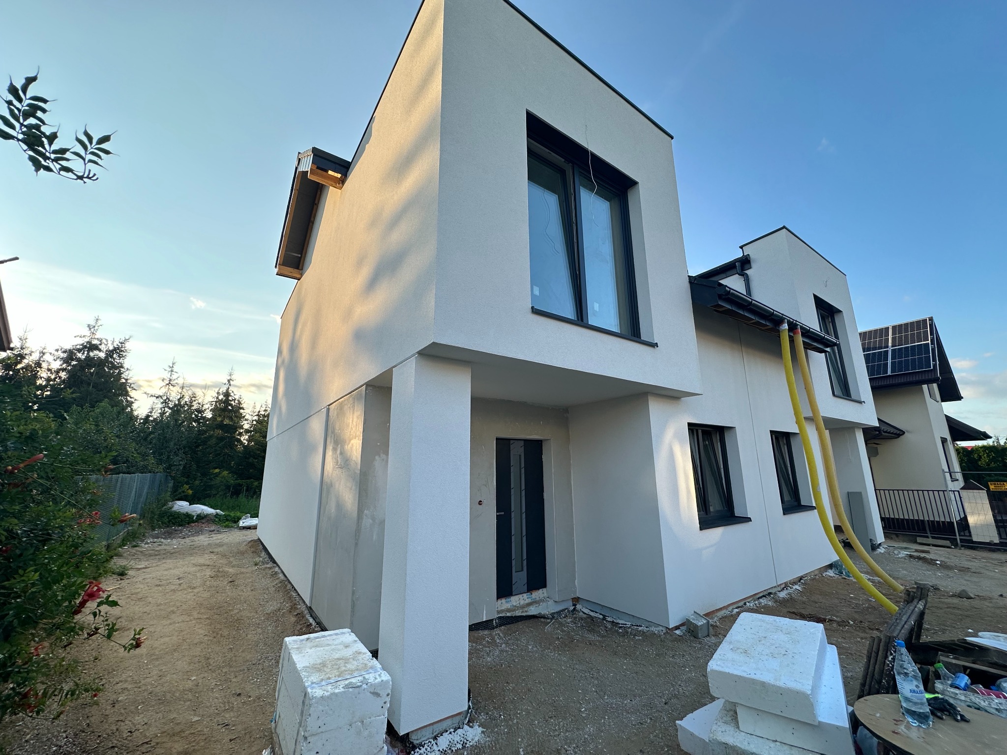 Nowoczesny dom z białą elewacją i czarnymi oknami, widoczny z zewnątrz. Część elewacji jest jeszcze surowa, co sugeruje trwające prace wykończeniowe.