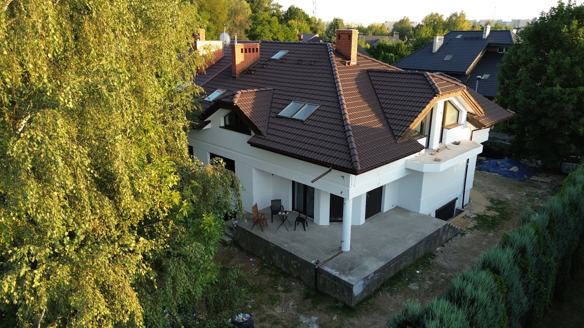 Biały dom z brązowym dachem w otoczeniu zieleni, widziany z góry. Widoczny taras z meblami ogrodowymi i fragment elewacji. Ujęcie w słoneczny dzień.