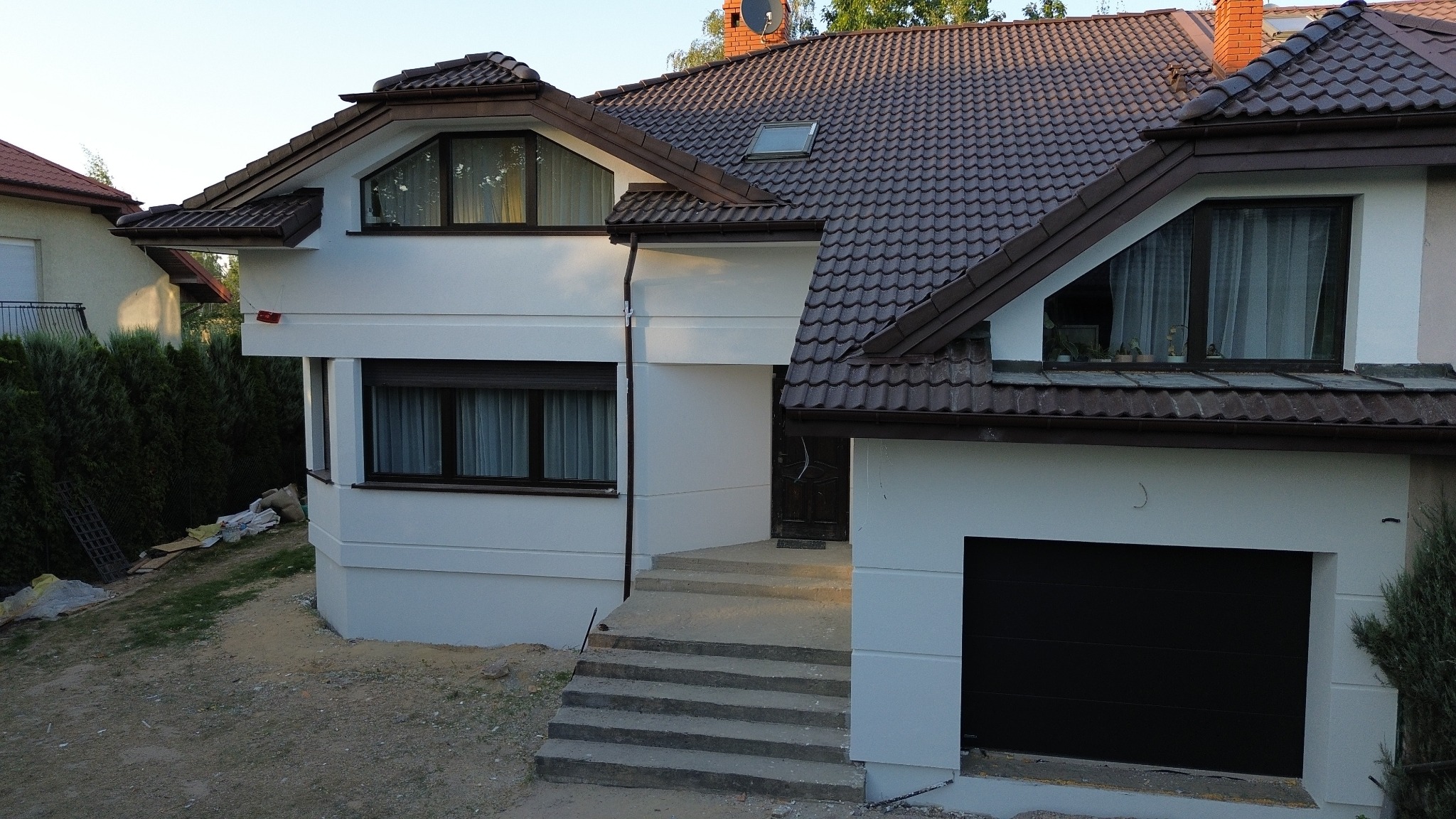 Biała elewacja domu jednorodzinnego z brązowym dachem i oknami. Widoczne schody wejściowe i brama garażowa. Częściowo zagospodarowany teren wokół budynku.