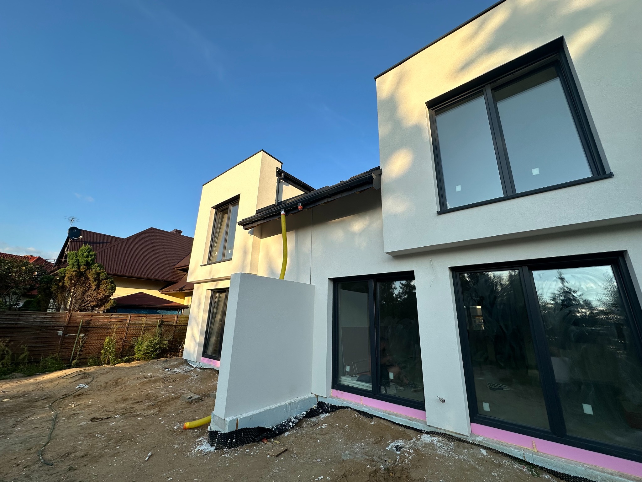 Nowoczesny dom z jasną elewacją i czarnymi ramami okiennymi, widok z dołu na tle błękitnego nieba. Wokół budynku widoczny teren w trakcie prac wykończeniowych.