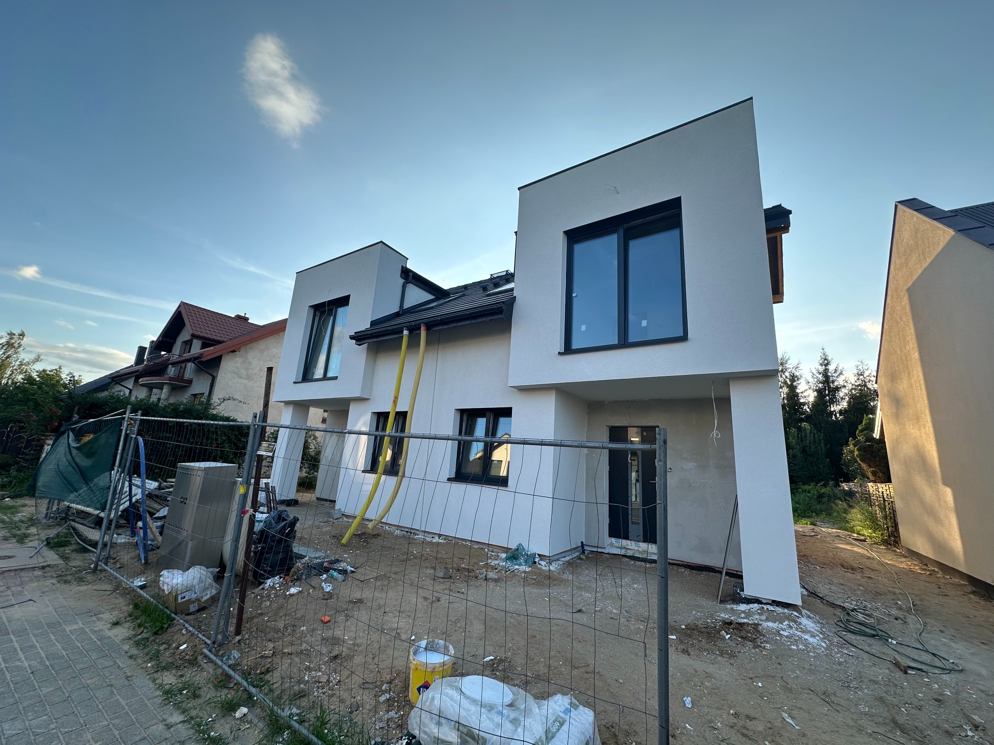 Nowoczesny, dwukondygnacyjny dom z białymi elewacjami i czarnymi ramami okiennymi, widziany z poziomu gruntu, z widocznym ogrodzeniem budowlanym i elementami wykończeniowymi.