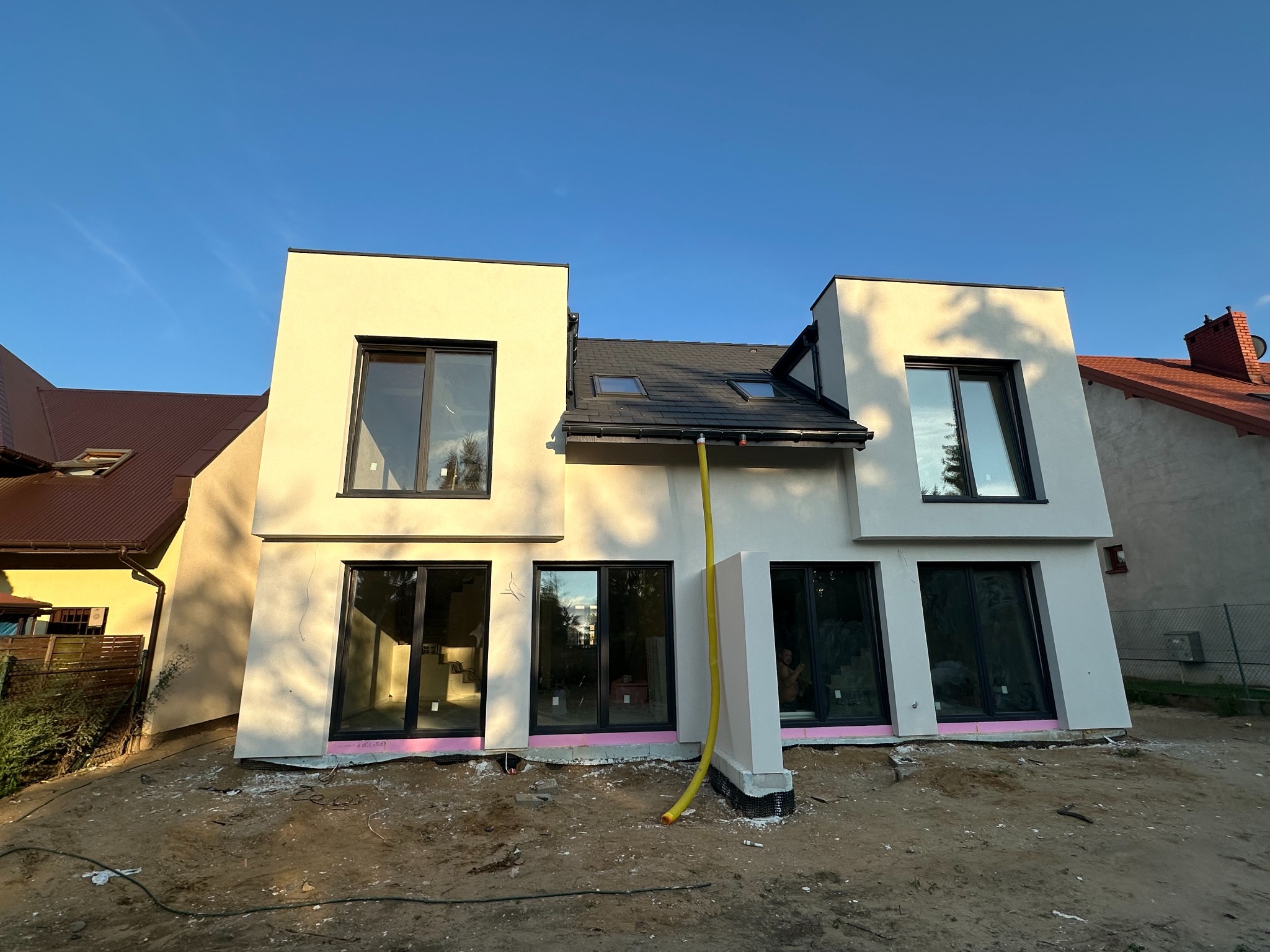 Nowoczesny dom z jasną elewacją, czarnymi ramami okiennymi i różowym ociepleniem fundamentów. Żółty wąż odprowadzający wodę. Błękitne niebo w tle.