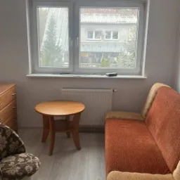 Jasne wnętrze pokoju z drewnianym stolikiem, brązową kanapą i krzesłem w kwiaty na podłodze z paneli, widok z okna na budynek z szarą dachówką.