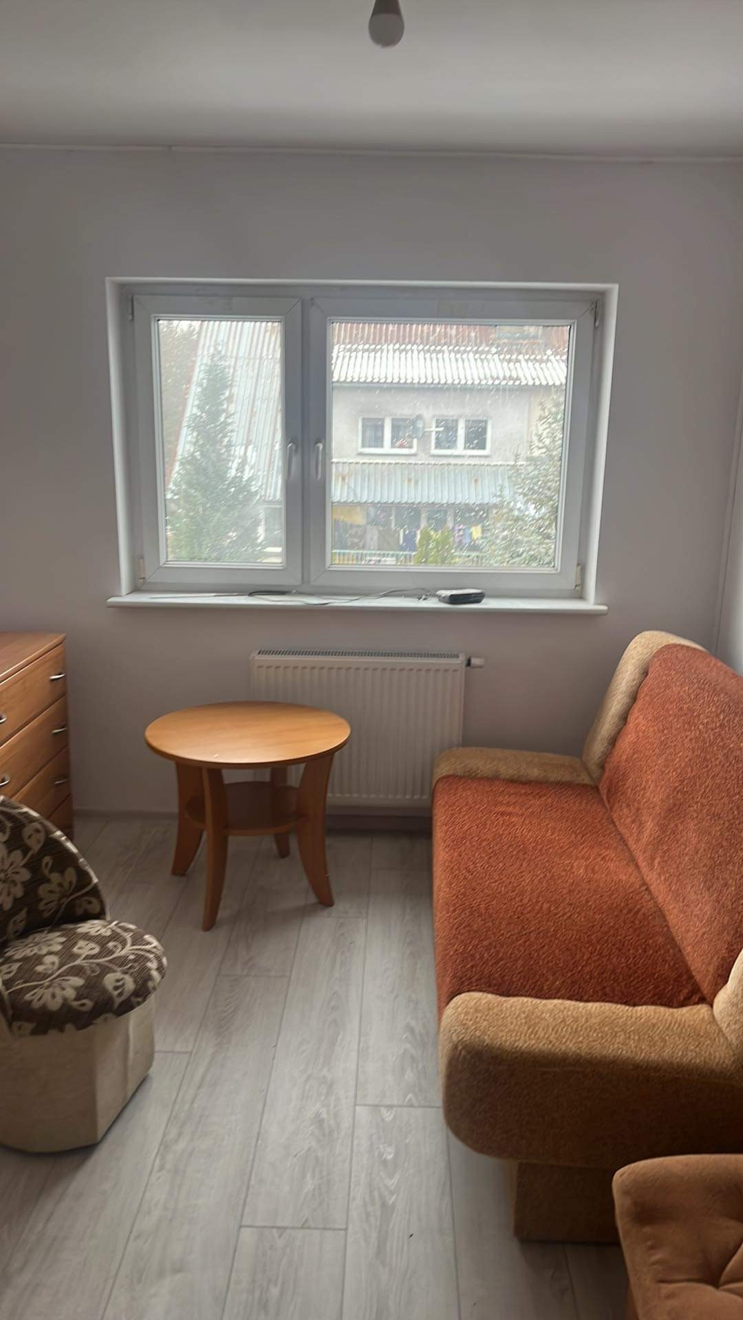 Jasne wnętrze pokoju z drewnianym stolikiem, brązową kanapą i krzesłem w kwiaty na podłodze z paneli, widok z okna na budynek z szarą dachówką.