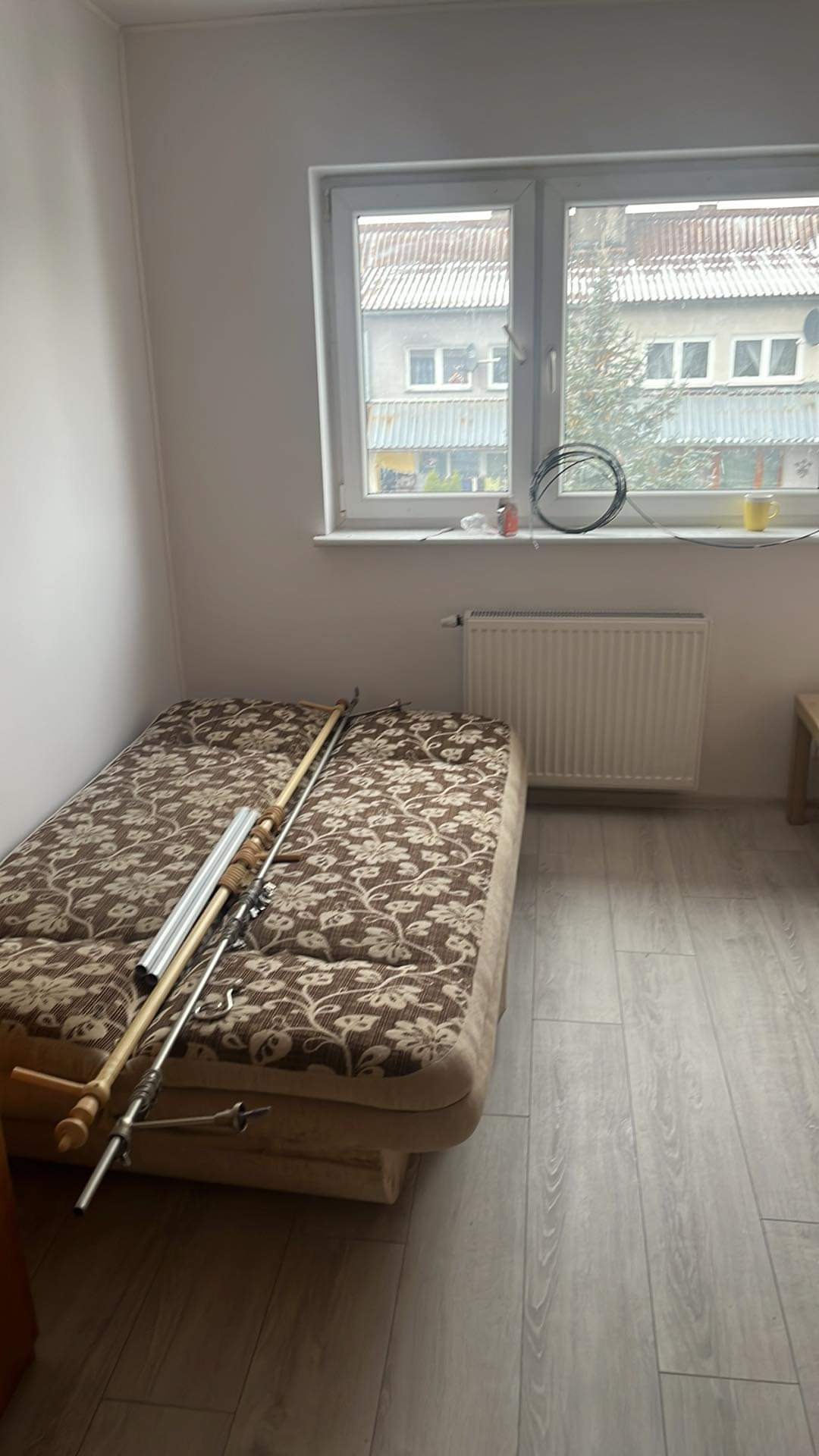 Pokój w trakcie przygotowań do remontu: widoczny rozłożony tapczan z narzędziami malarskimi, grzejnik pod oknem, panele podłogowe i ściany przygotowane do malowania.