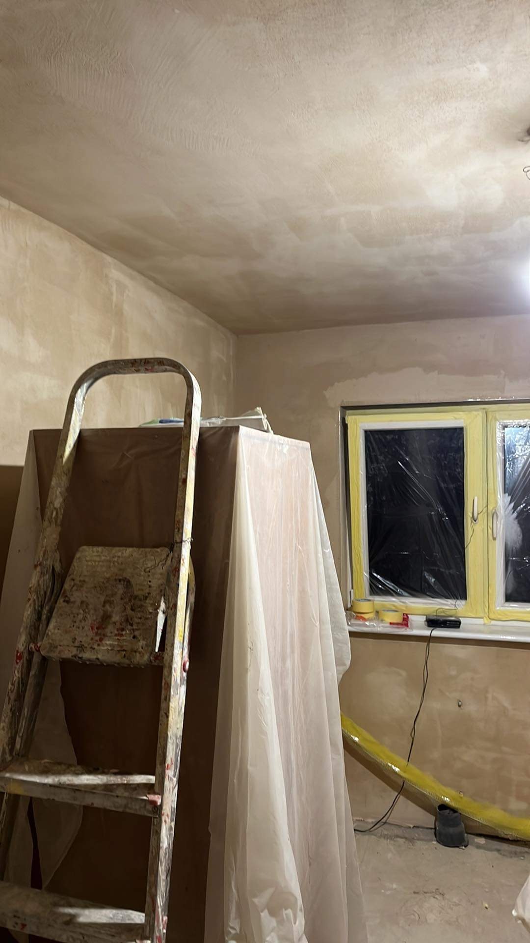 Pokój w trakcie prac wykończeniowych: ściany i sufit po nałożeniu gładzi, okno zabezpieczone folią, drabina malarska z resztkami farby i meble przykryte folią ochronną.