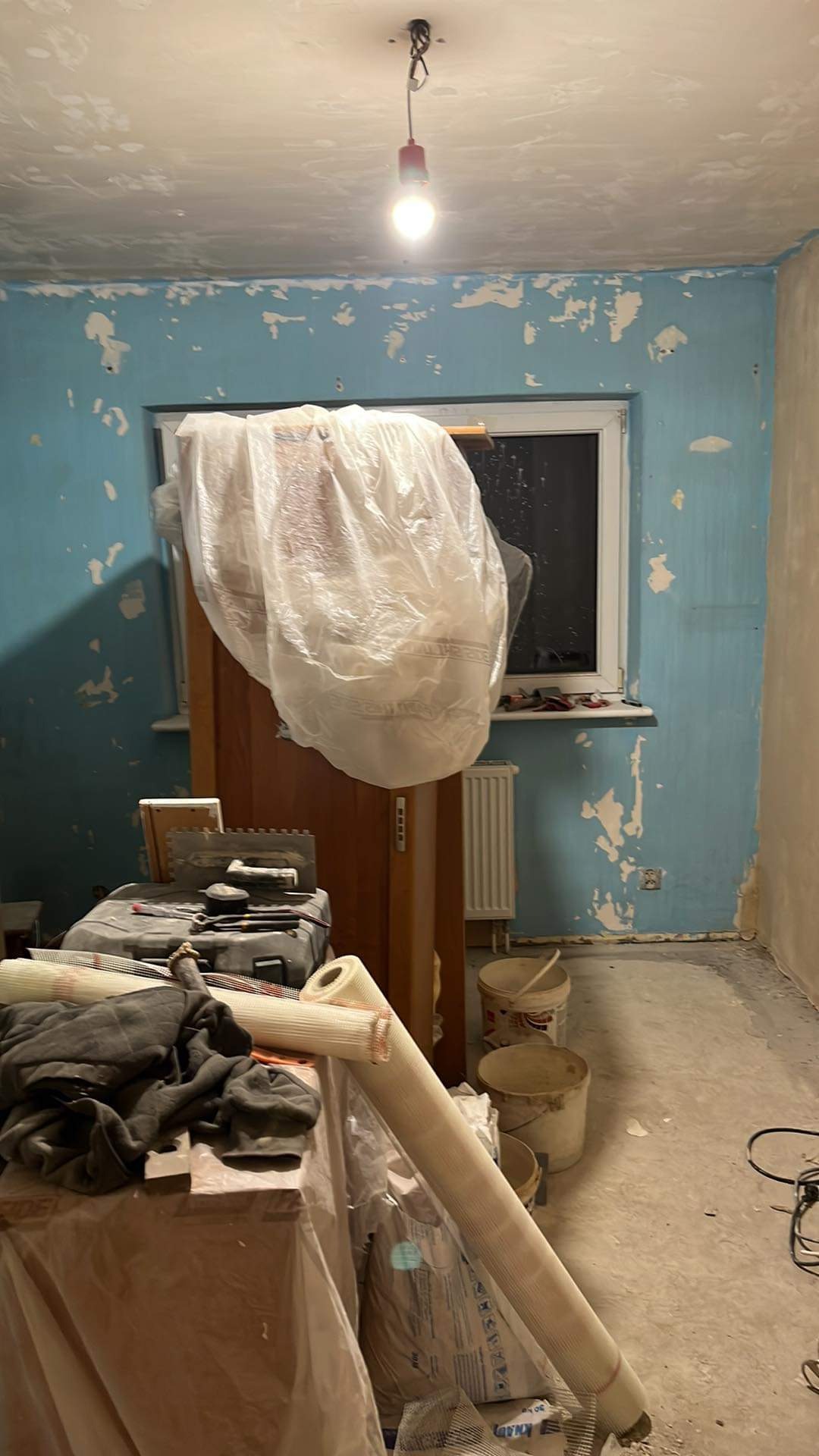 Pokój w trakcie przygotowania do prac wykończeniowych: ściany z resztkami farby, okno, grzejnik, zakryte meble, narzędzia i materiały budowlane w nieładzie, oświetlenie żarówką.