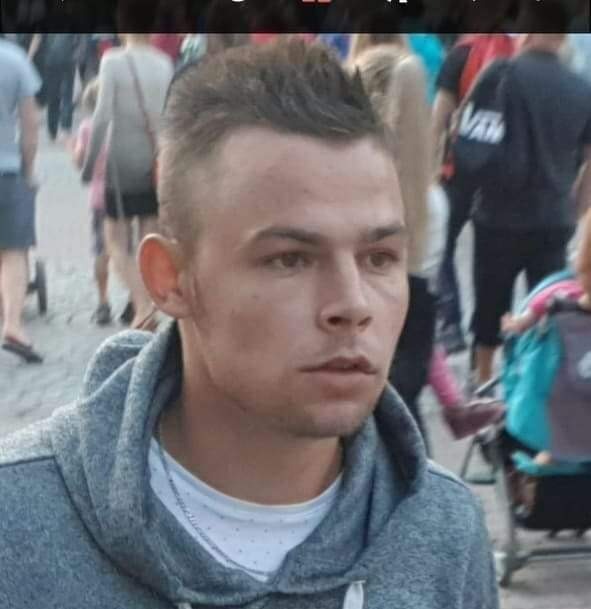 Portret młodego mężczyzny w szarej bluzie z kapturem na tle rozmytego tłumu w plenerze.