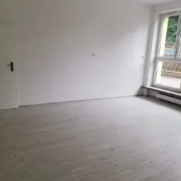 Pusty pokój po remoncie z nowymi panelami podłogowymi w odcieniach szarości i świeżo pomalowanymi na biało ścianami, widoczny grzejnik pod oknem.