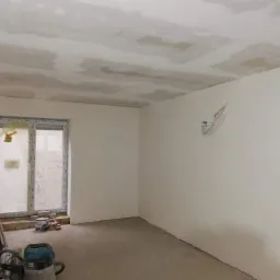 Pokój w trakcie prac wykończeniowych: sufit z widocznymi łatami po szpachlowaniu, ściany przygotowane pod malowanie, widoczne przewody elektryczne, okno zabezpieczone folią.