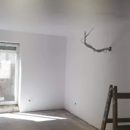 Wnętrze pokoju po świeżym pomalowaniu na biało, widoczne ślady po pracach wykończeniowych, drabina oparta o ścianę, wiadro z farbą i wałki malarskie na podłodze, surowe przewody wystające ze ściany.