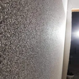 Szara ściana pokryta dekoracyjną, ziarnistą strukturą, z widocznym narożnikiem i fragmentem otworu drzwiowego w tle.