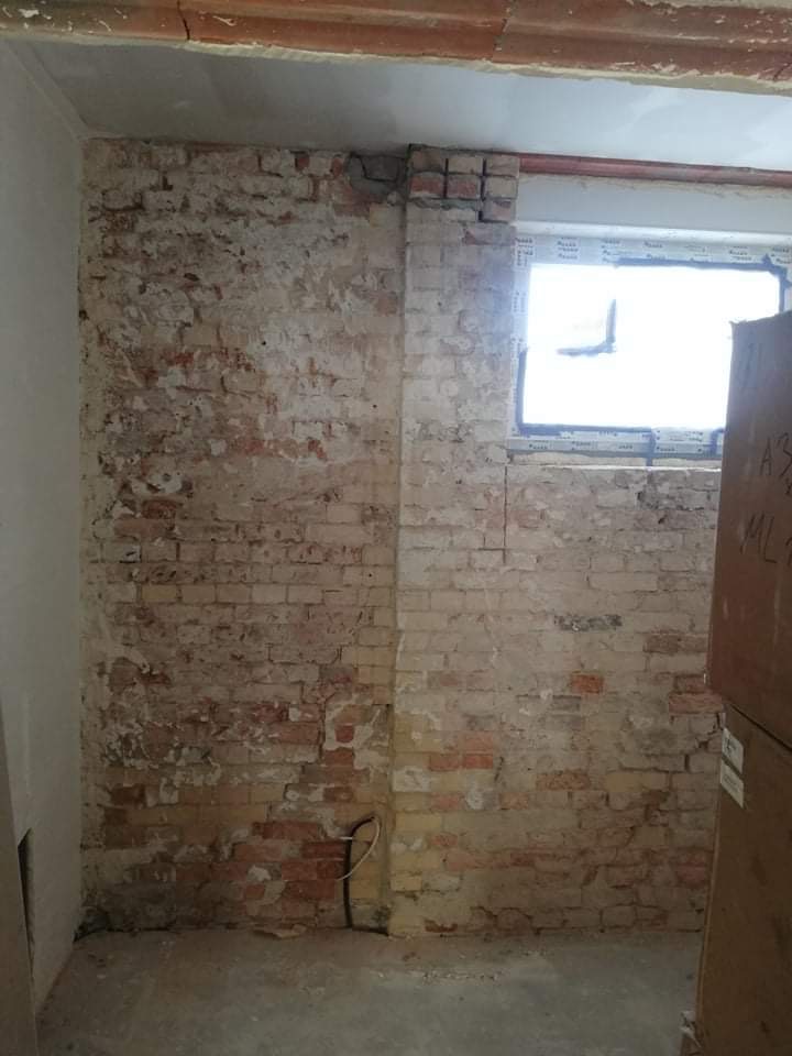 Surowa ściana z cegły w trakcie remontu, widoczne pozostałości tynku, okno z plastikową ramą i wystający kabel elektryczny.