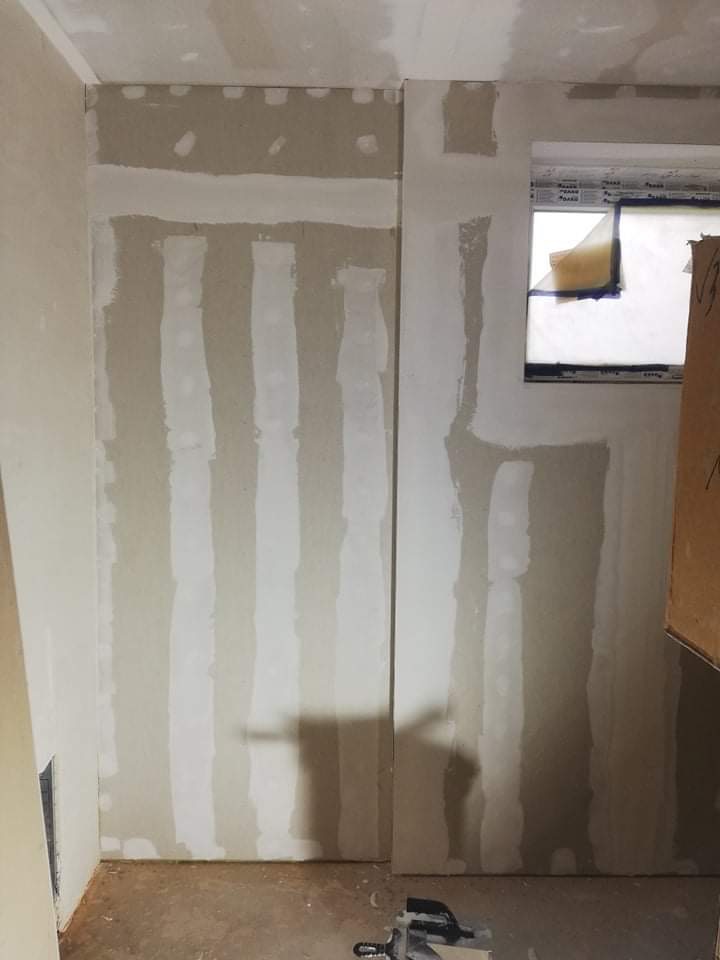 Ściana w trakcie prac przygotowawczych do wykończenia: pionowe pasy nałożonej warstwy wyrównującej, surowy stan podłoża i okno zabezpieczone folią malarską.