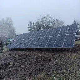 Ustawiony na gruncie panel fotowoltaiczny w zimowej scenerii, z lekką mgłą i oszronionymi drzewami w tle, obok zaparkowany bus.