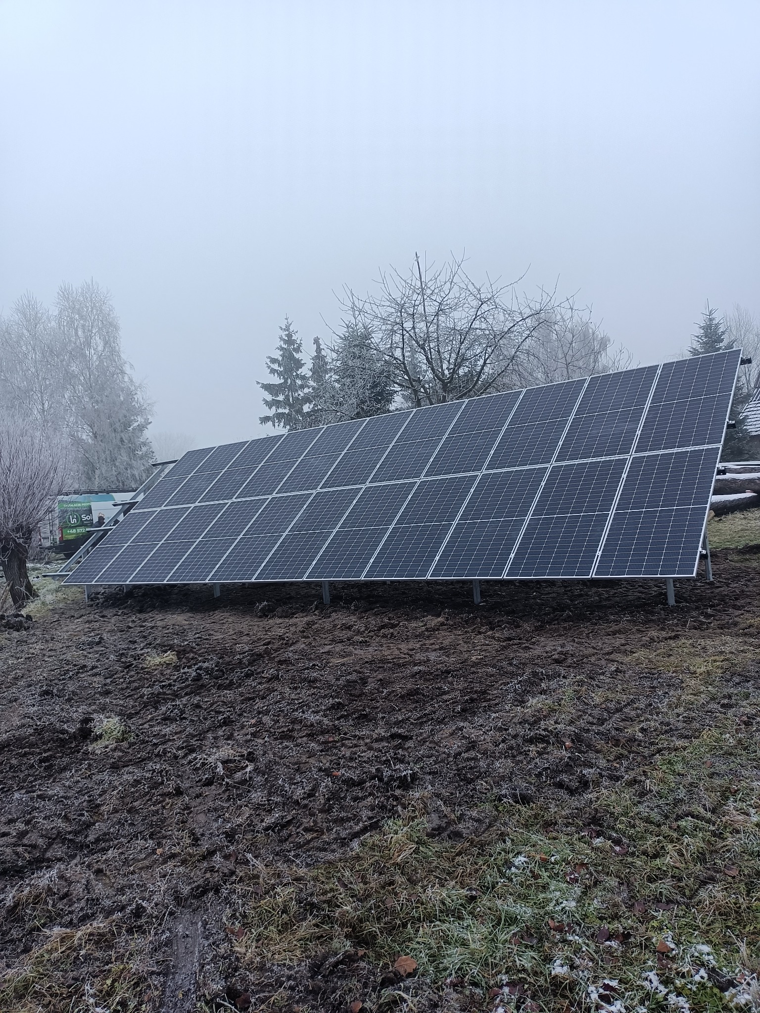 Ustawiony na gruncie panel fotowoltaiczny w zimowej scenerii, z lekką mgłą i oszronionymi drzewami w tle, obok zaparkowany bus.