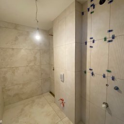 Michał Worobiej - Świeżo ułożone, beżowe płytki w łazience, z widocznymi krzyżykami dystansowymi w kolorze niebieskim i zielonym, oczekujące na fugowanie. Wisząca żarówka oświetla wnętrze.