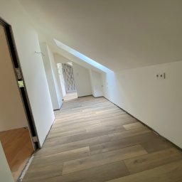 Michał Worobiej - Poddasze po remoncie: świeżo ułożone panele podłogowe w odcieniach beżu i brązu, białe ściany z widocznymi gniazdkami elektrycznymi, surowe ościeżnice drzwiowe i fragment łazienki w tle.