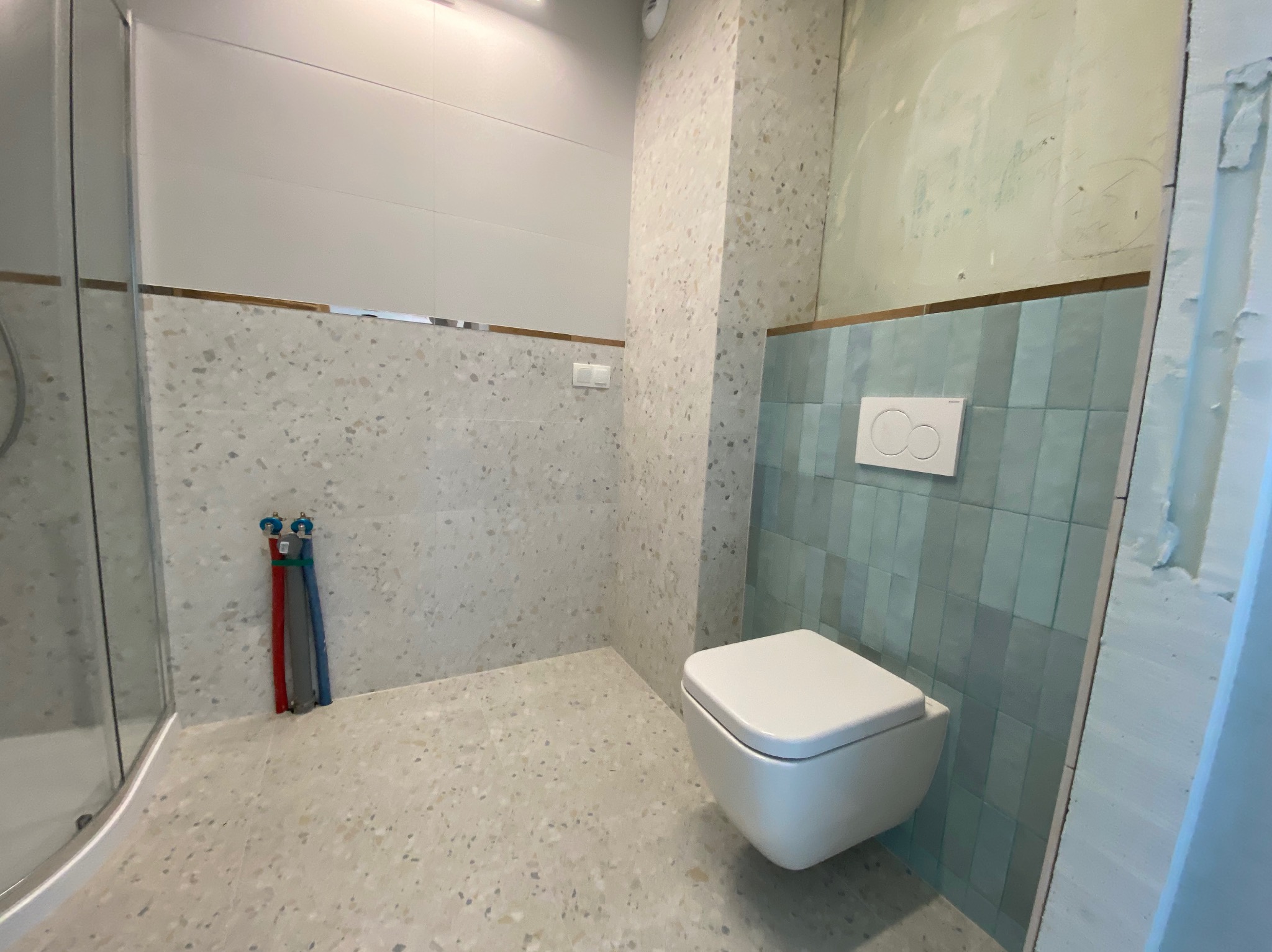 Wnętrze łazienki w trakcie remontu z wiszącą toaletą, niebieskimi płytkami na ścianie oraz widocznymi instalacjami hydraulicznymi i fragmentem kabiny prysznicowej.