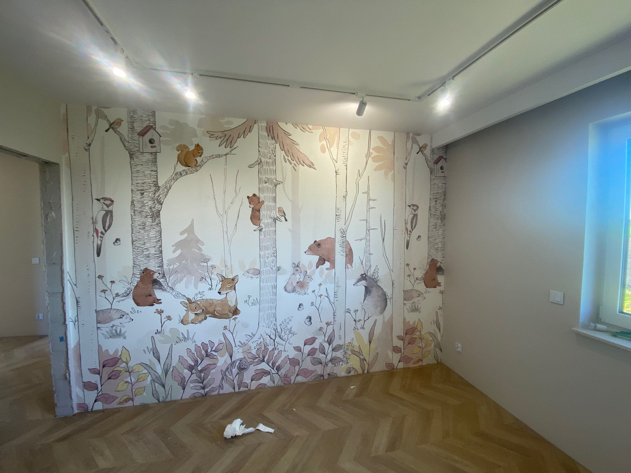 Pokój dziecięcy z fototapetą przedstawiającą leśne zwierzęta, drewniana podłoga w jodełkę, oświetlenie szynowe na suficie, widoczne ślady po remoncie.