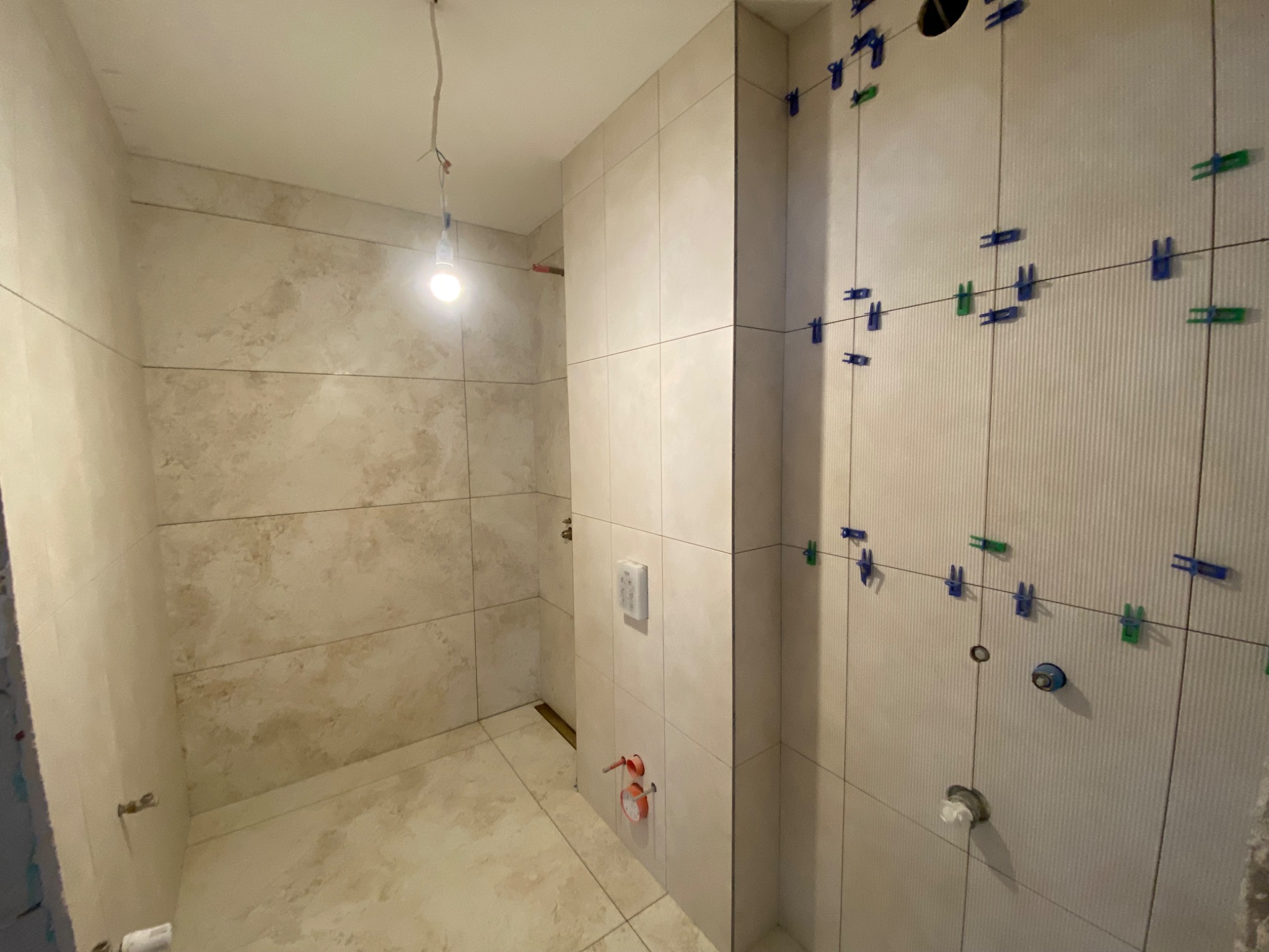 Świeżo ułożone, beżowe płytki w łazience, z widocznymi krzyżykami dystansowymi w kolorze niebieskim i zielonym, oczekujące na fugowanie. Wisząca żarówka oświetla wnętrze.