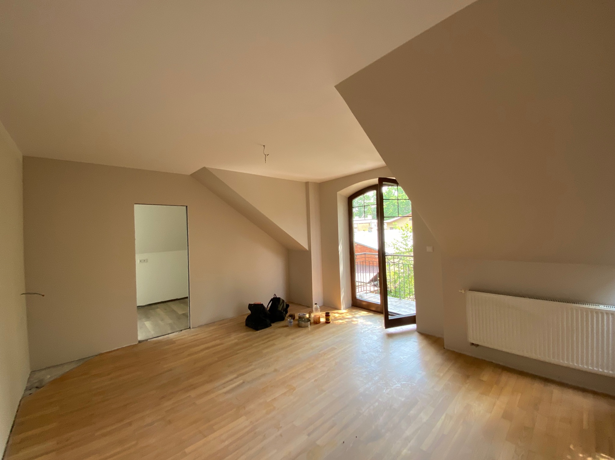 Pokój na poddaszu po malowaniu ścian na beżowo, z widocznym fragmentem podłogi przed wykończeniem, otwartym oknem balkonowym i grzejnikiem.