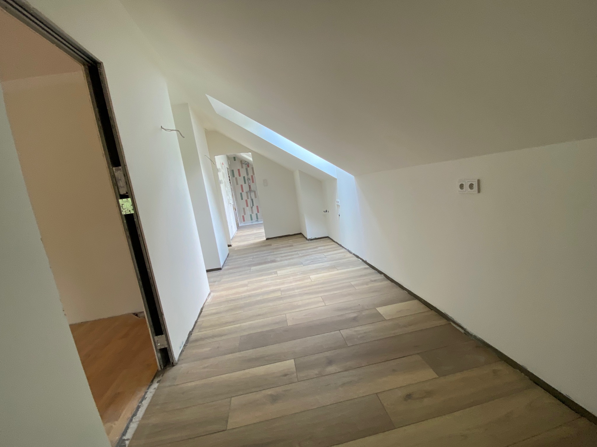 Poddasze po remoncie: świeżo ułożone panele podłogowe w odcieniach beżu i brązu, białe ściany z widocznymi gniazdkami elektrycznymi, surowe ościeżnice drzwiowe i fragment łazienki w tle.