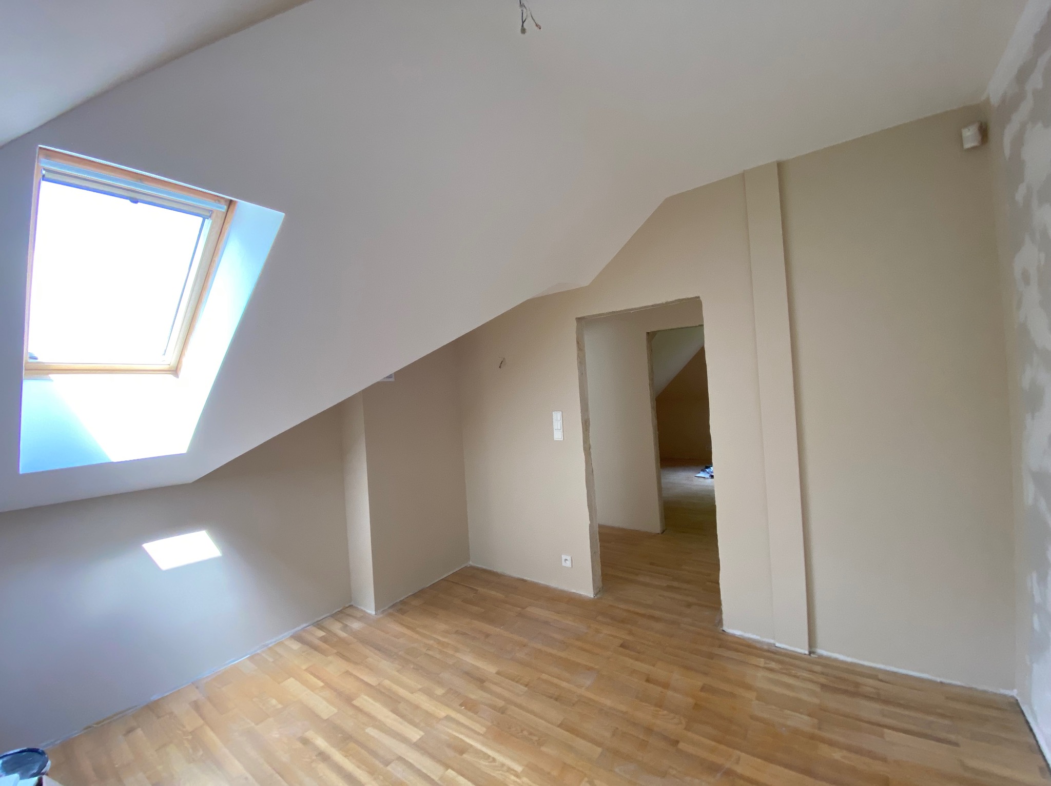 Poddasze po malowaniu ścian na beżowo, z drewnianą podłogą i oknem dachowym, widoczne ślady po remoncie przy listwach przypodłogowych.