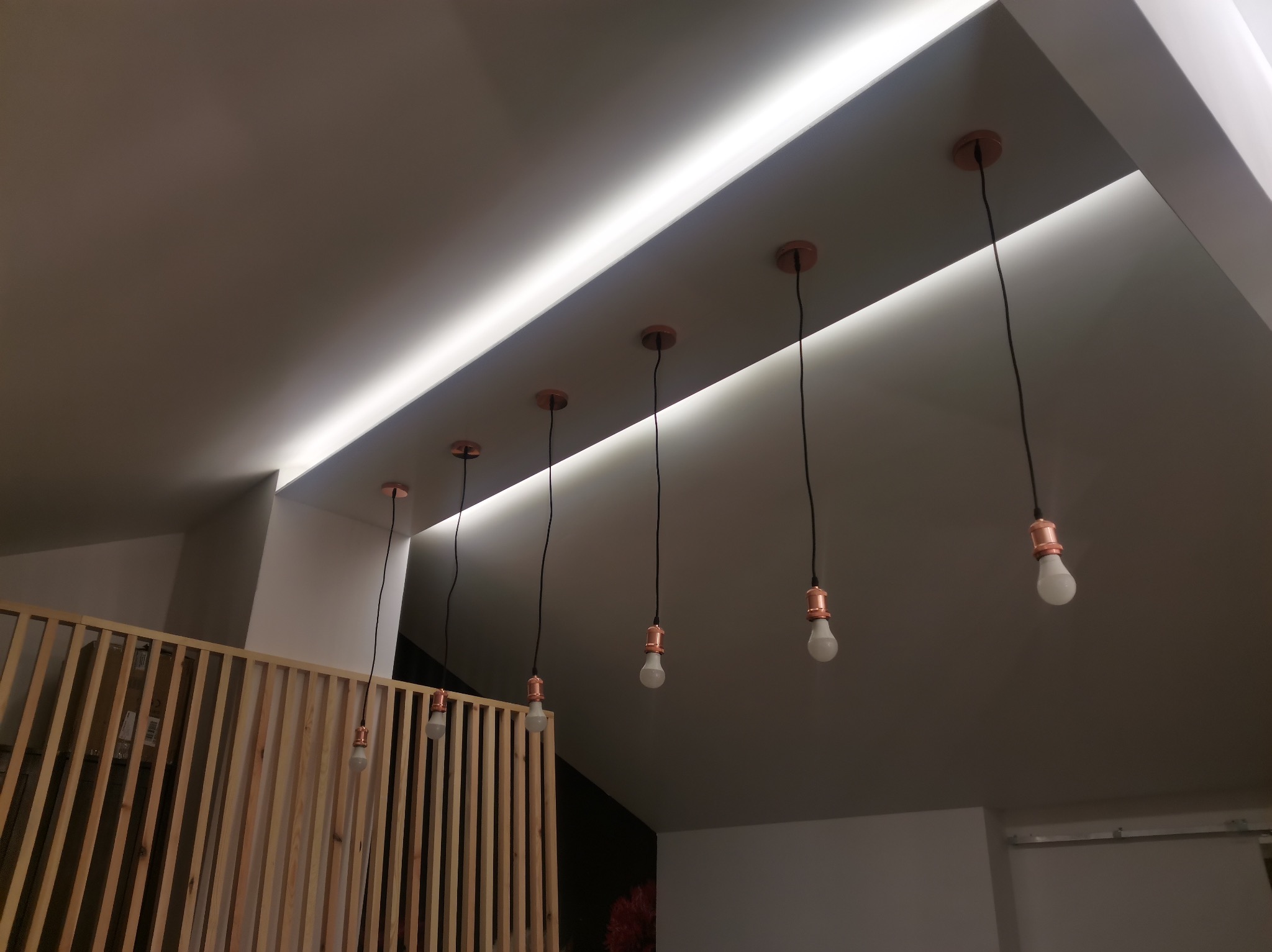 Biały sufit z wbudowanym oświetleniem LED i rzędem wiszących żarówek z miedzianymi oprawkami, widoczna drewniana ażurowa ścianka działowa.