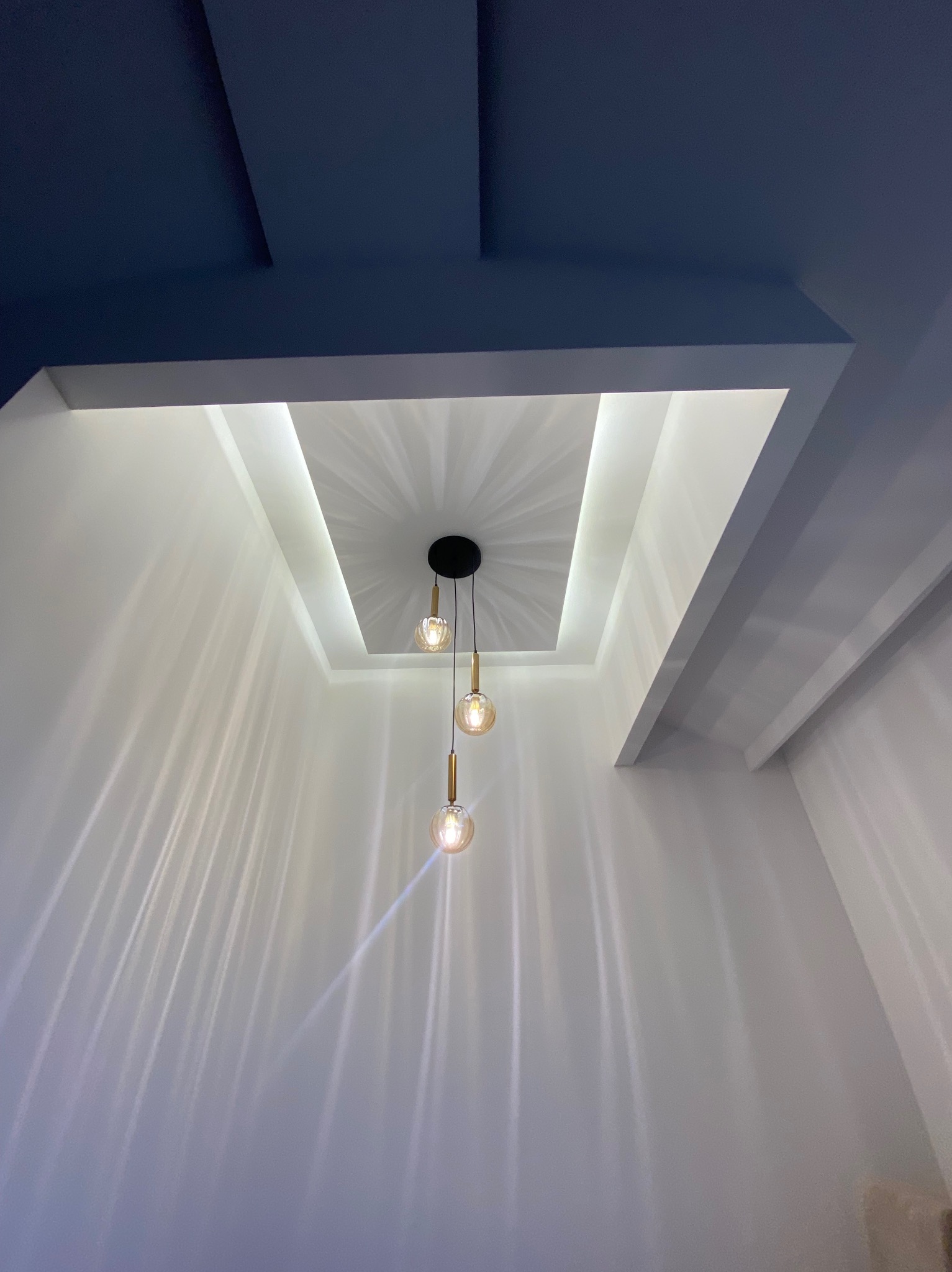 Nowoczesny sufit podwieszany z oświetleniem LED w kształcie prostokąta i trzema wiszącymi lampami z żarówkami dekoracyjnymi, rzucającymi długie cienie na białe ściany.