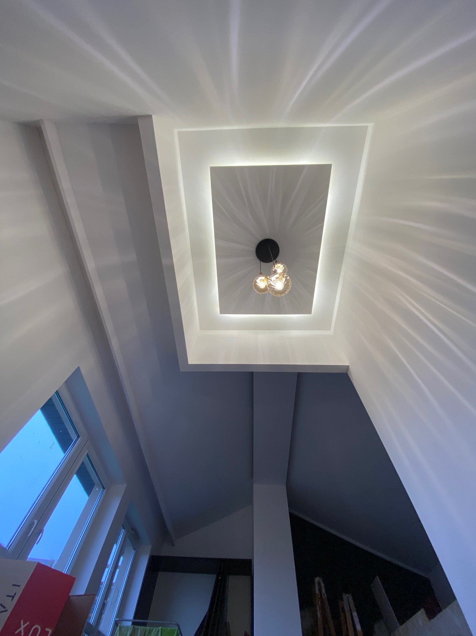 Biały sufit z wnęką oświetleniową i nowoczesną lampą wiszącą z trzema szklanymi kulami, światło odbija się od ścian