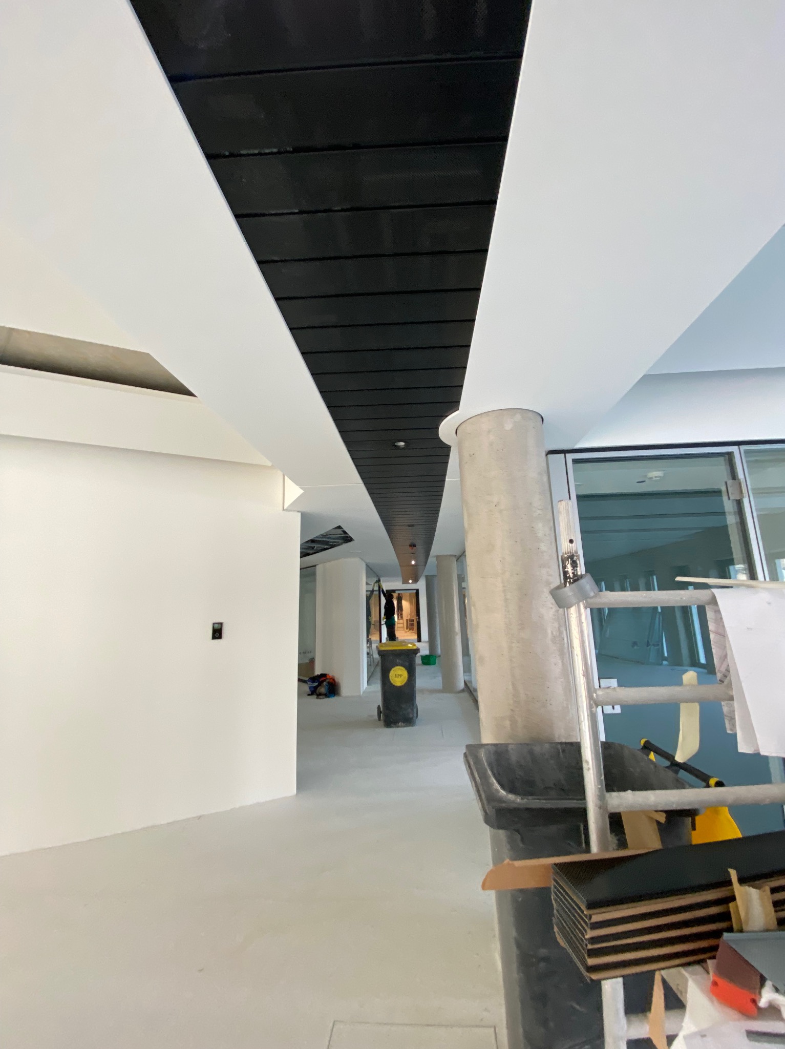 Wnętrze w trakcie prac wykończeniowych: widoczny sufit z czarnymi panelami i białymi pasami, surowy betonowy filar, drabina, śmieci i narzędzia malarskie w tle, sugerujące etap prac w obiekcie...