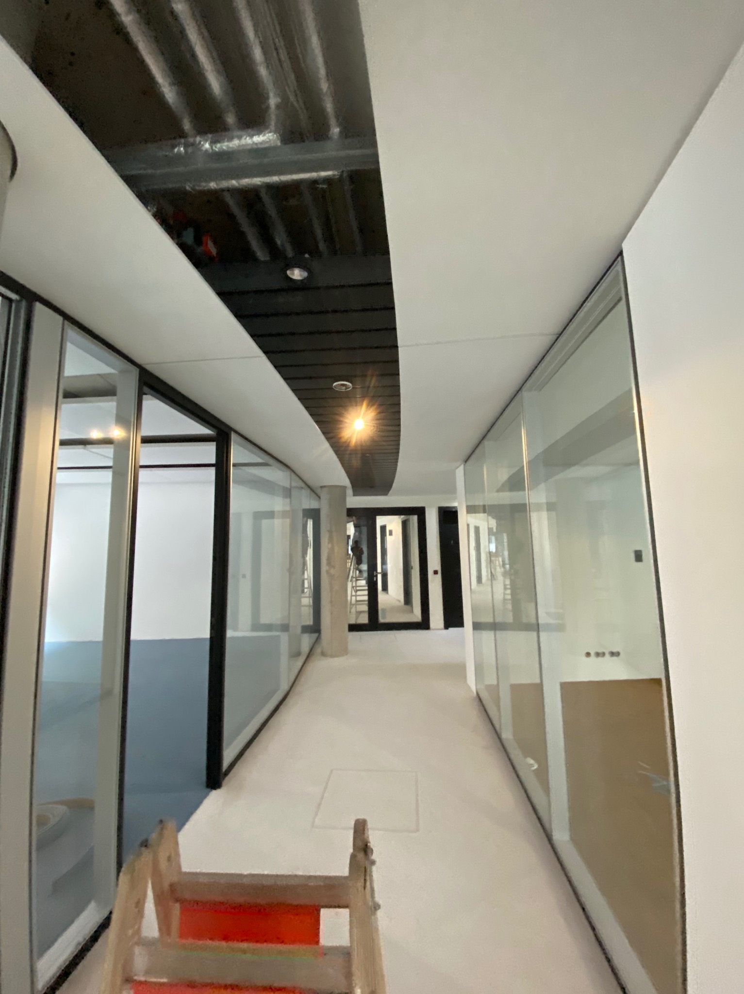 Korytarz biurowy w trakcie prac wykończeniowych, widoczna surowa konstrukcja sufitu, szklane ścianki działowe i drewniana drabina malarska.