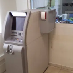 Szary bankomat z rolką papieru termicznego obok, umieszczony w pomieszczeniu z oknem i kafelkami na podłodze.