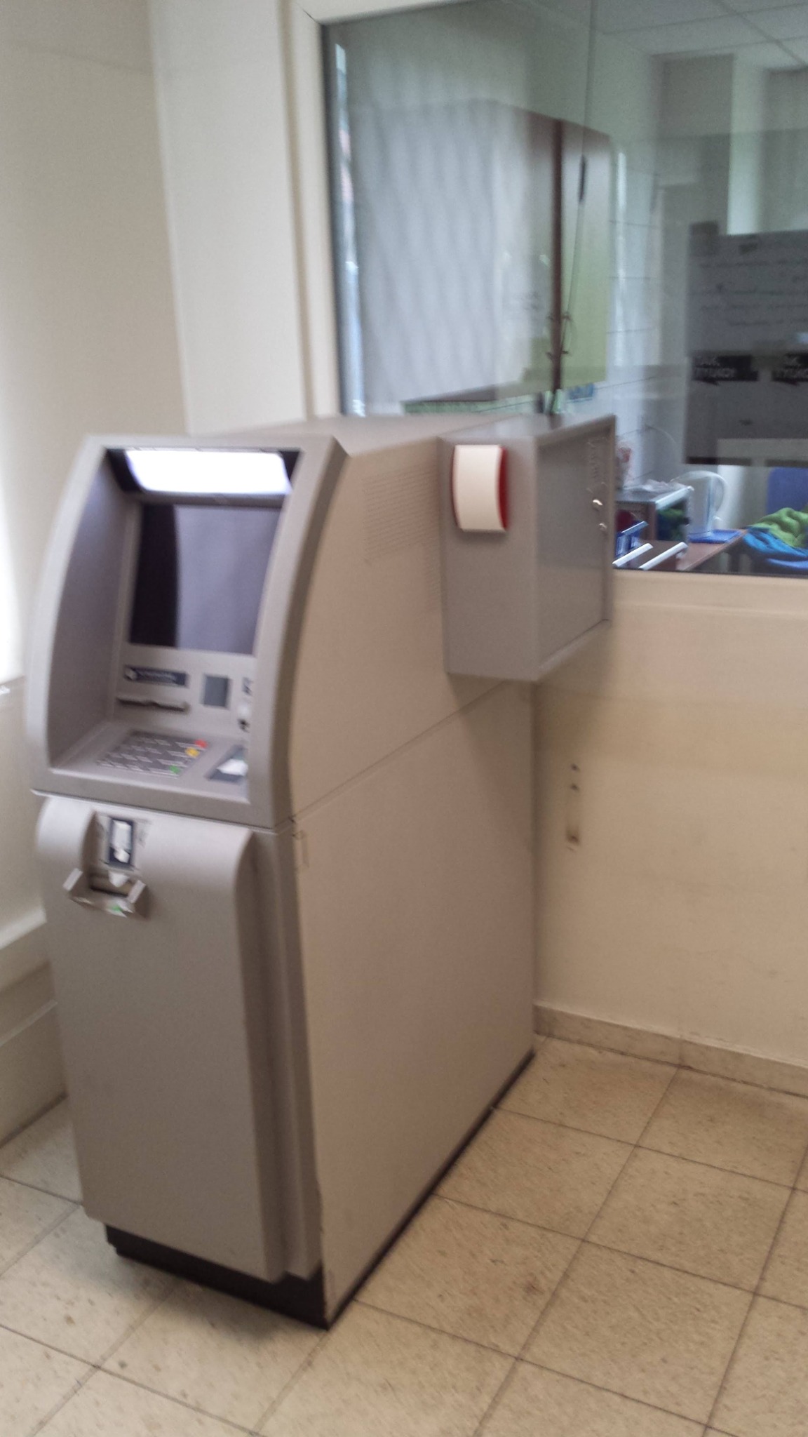 Szary bankomat z rolką papieru termicznego obok, umieszczony w pomieszczeniu z oknem i kafelkami na podłodze.