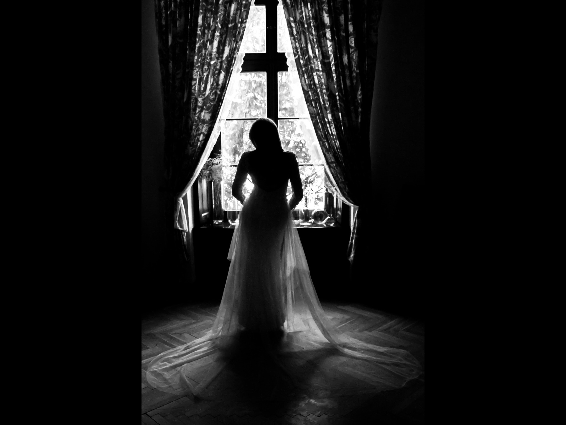 Czarno-białe zdjęcie sylwetki panny młodej w sukni ślubnej stojącej tyłem do okna z zasłonami w ciemnym pokoju.