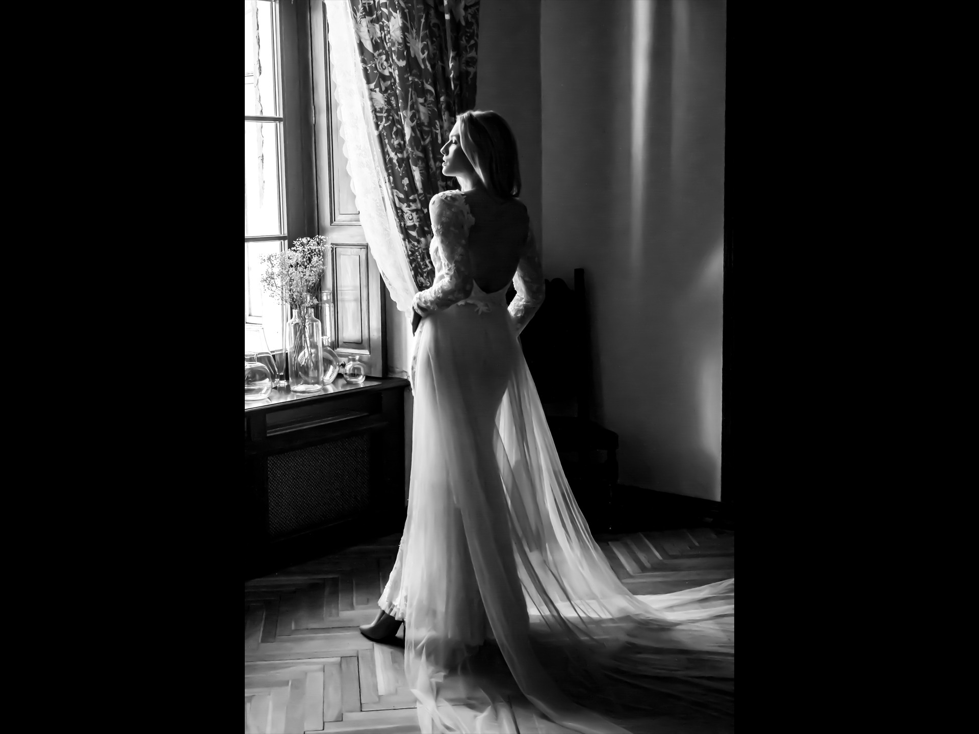 Czarno-białe zdjęcie kobiety w sukni z odkrytymi plecami, stojącej tyłem przy oknie z kwiatami w wazonach, na tle wzorzystej zasłony i drewnianej podłogi.