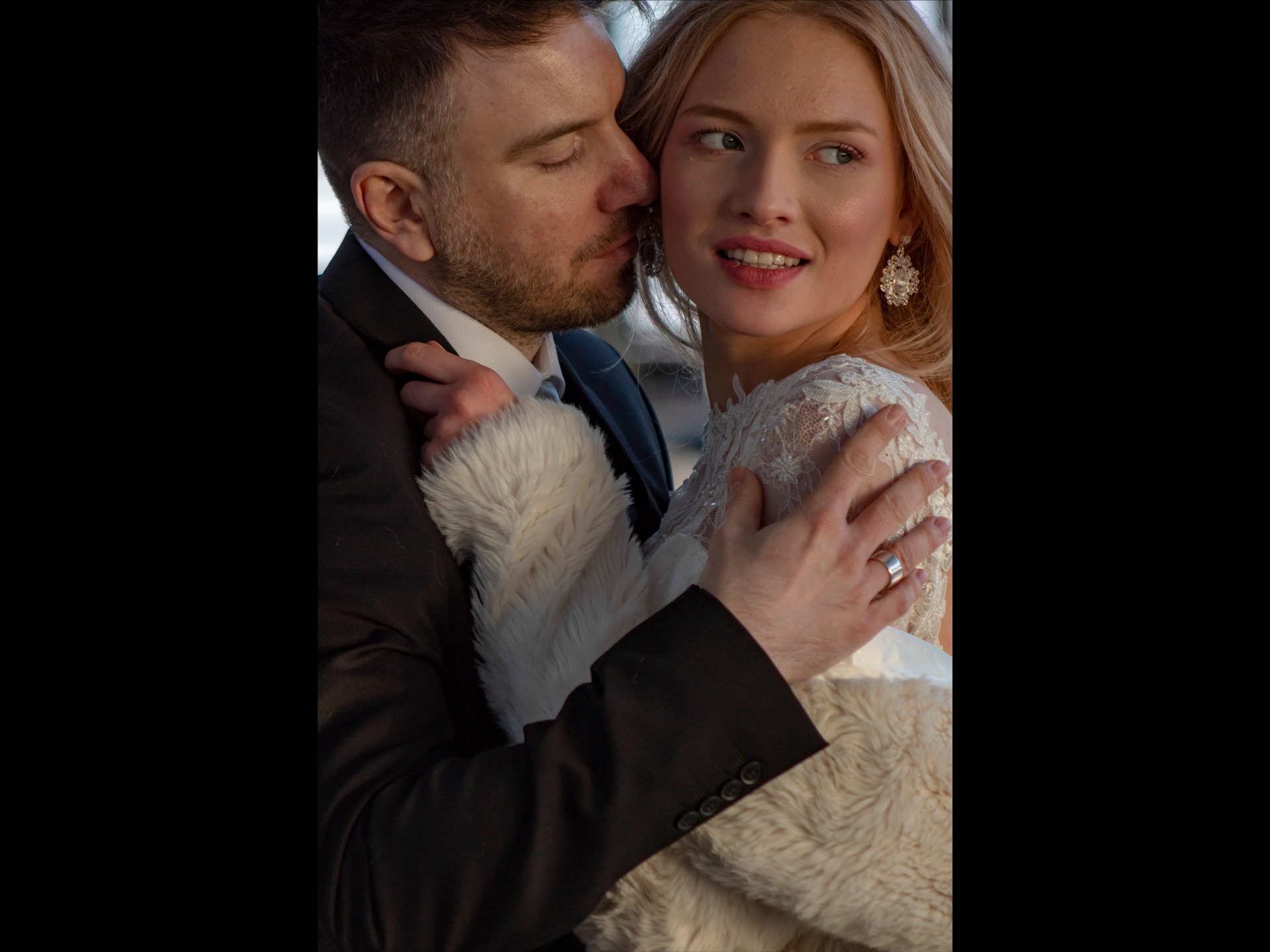 Mężczyzna w garniturze całuje w policzek uśmiechniętą blondynkę w koronkowej sukni i futrzanym okryciu.