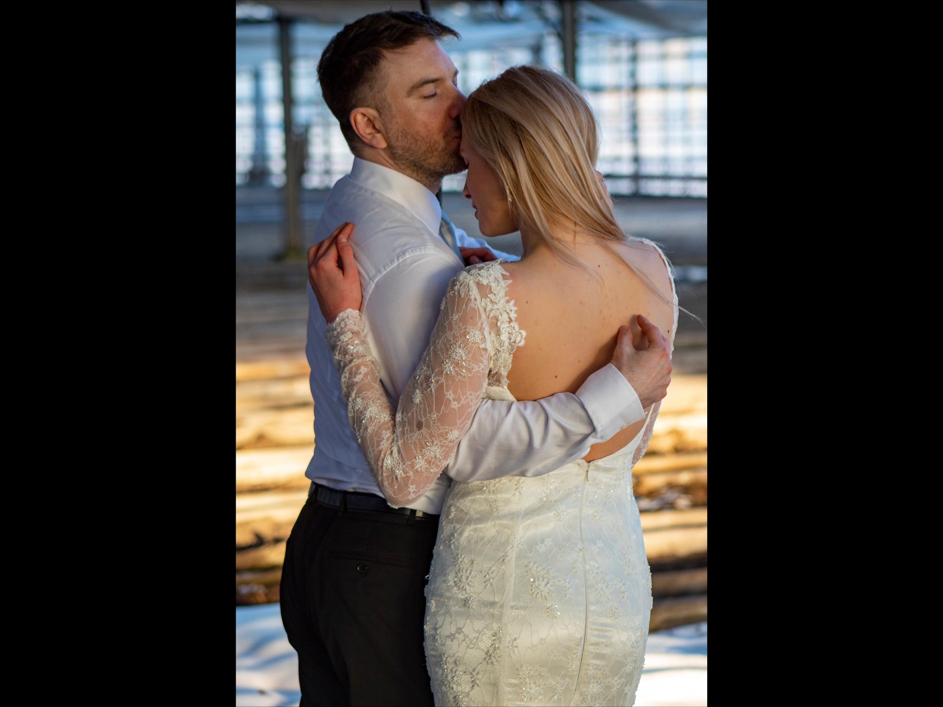 Nowożeńcy w objęciach: mężczyzna całuje kobietę w czoło, ona w sukni z koronkowymi rękawami, tło rozmyte.