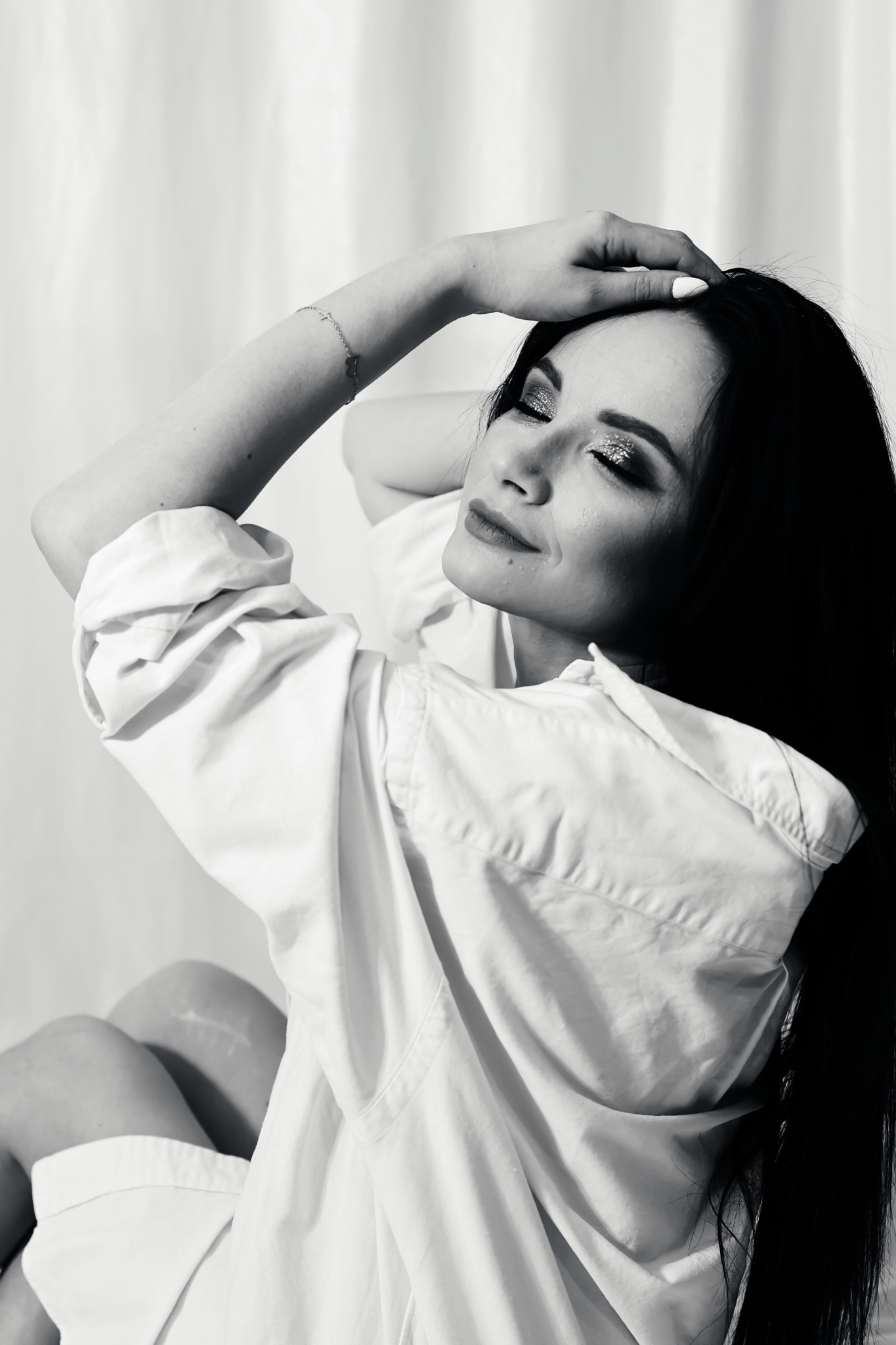 Czarno-białe zdjęcie kobiety z długimi, ciemnymi włosami, w białej koszuli, z błyszczącym makijażem oczu, z ręką na głowie i delikatną bransoletką na nadgarstku.