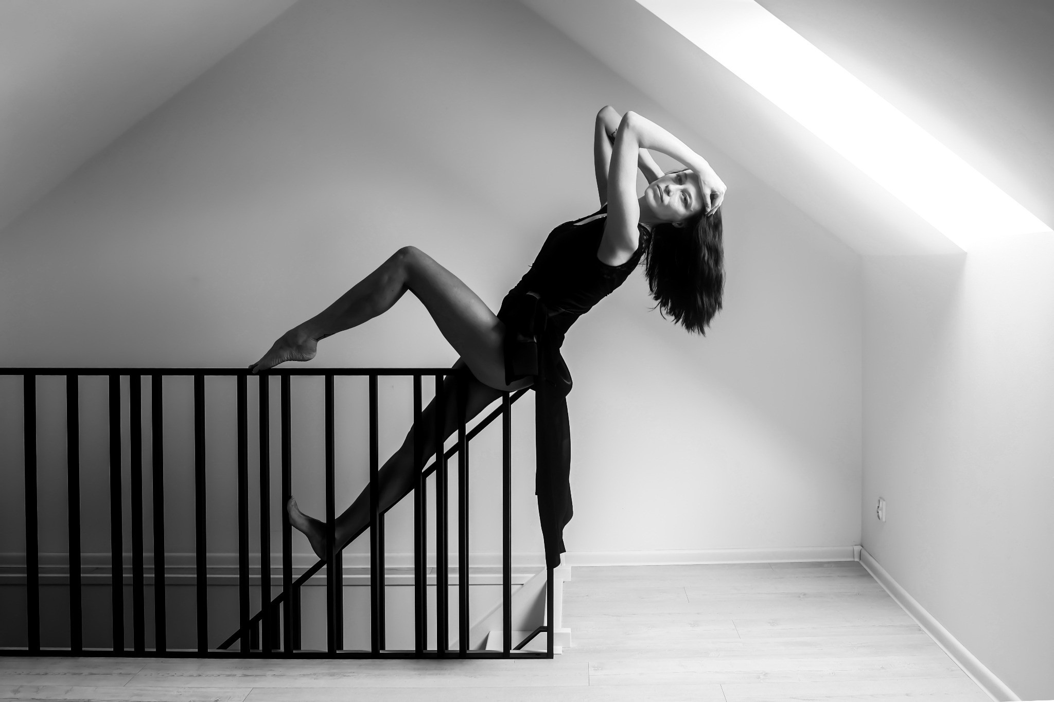 Kobieta w czarnym stroju pozuje na schodach z metalową barierką w minimalistycznym wnętrzu, czarno-biała fotografia z mocnym kontrastem, nietypowa perspektywa.