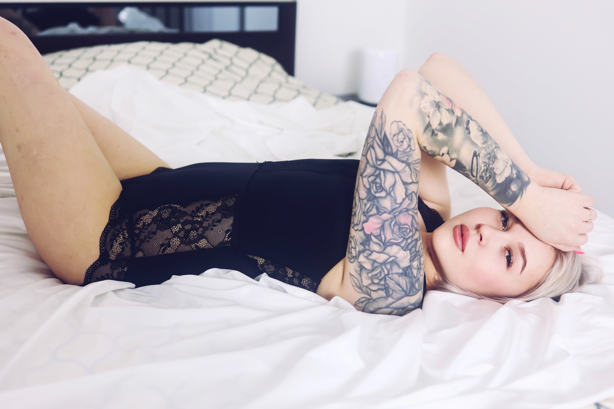 Kobieta z tatuażami na ramionach w czarnym body z koronkowymi wstawkami leży na białej pościeli, spoglądając w obiektyw.