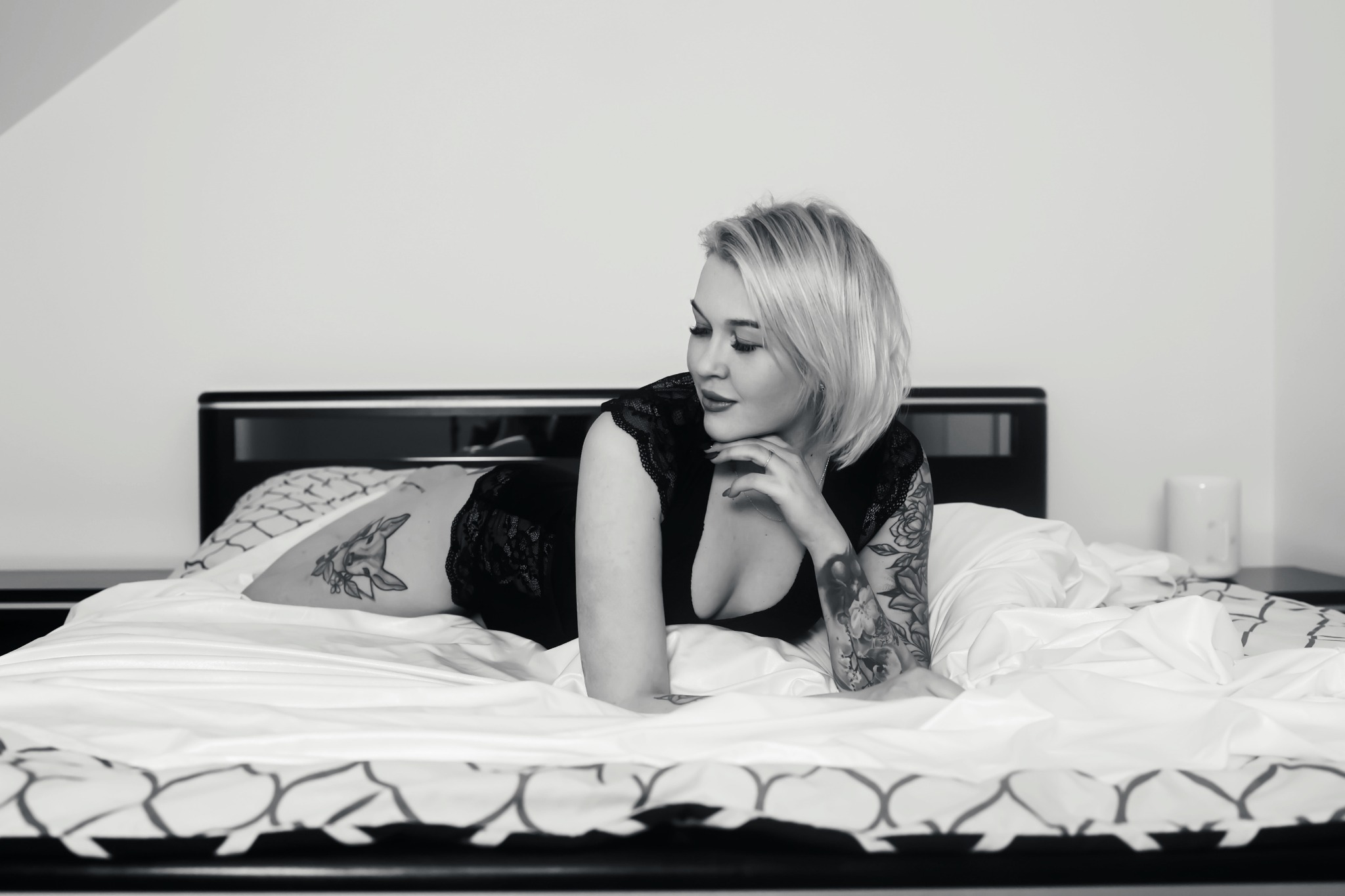 Kobieta z tatuażami, w koronkowej bieliźnie leży na łóżku, z głową opartą na dłoni, spoglądając w bok.