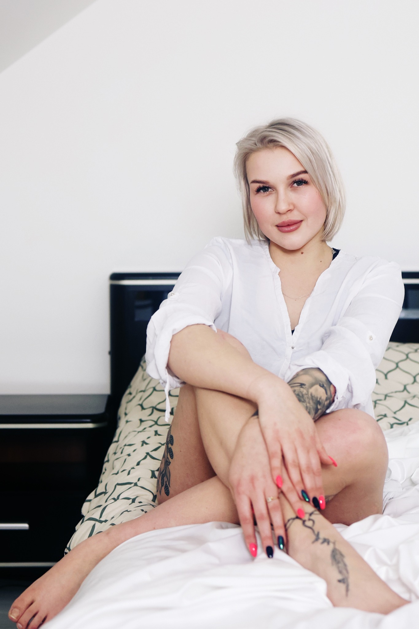 Portret kobiety z platynowymi włosami, w białej koszuli, siedzącej na łóżku z wzorzystą pościelą, z tatuażami na rękach i nogach, prezentującej manicure hybrydowy w odcieniach różu i czerni.