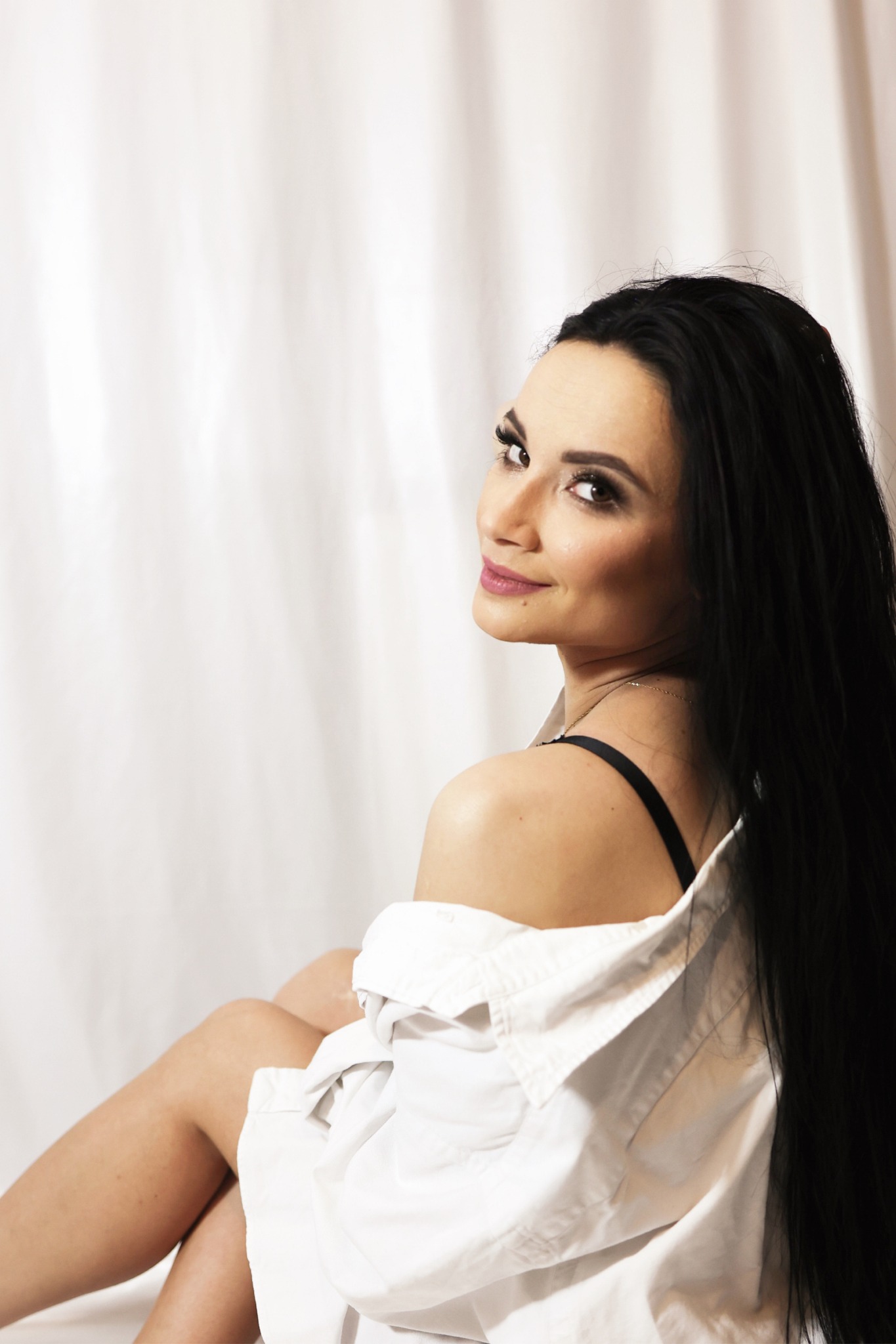 Portret kobiety z długimi czarnymi włosami, odwróconej przez ramię, w białej koszuli na tle białej draperii.