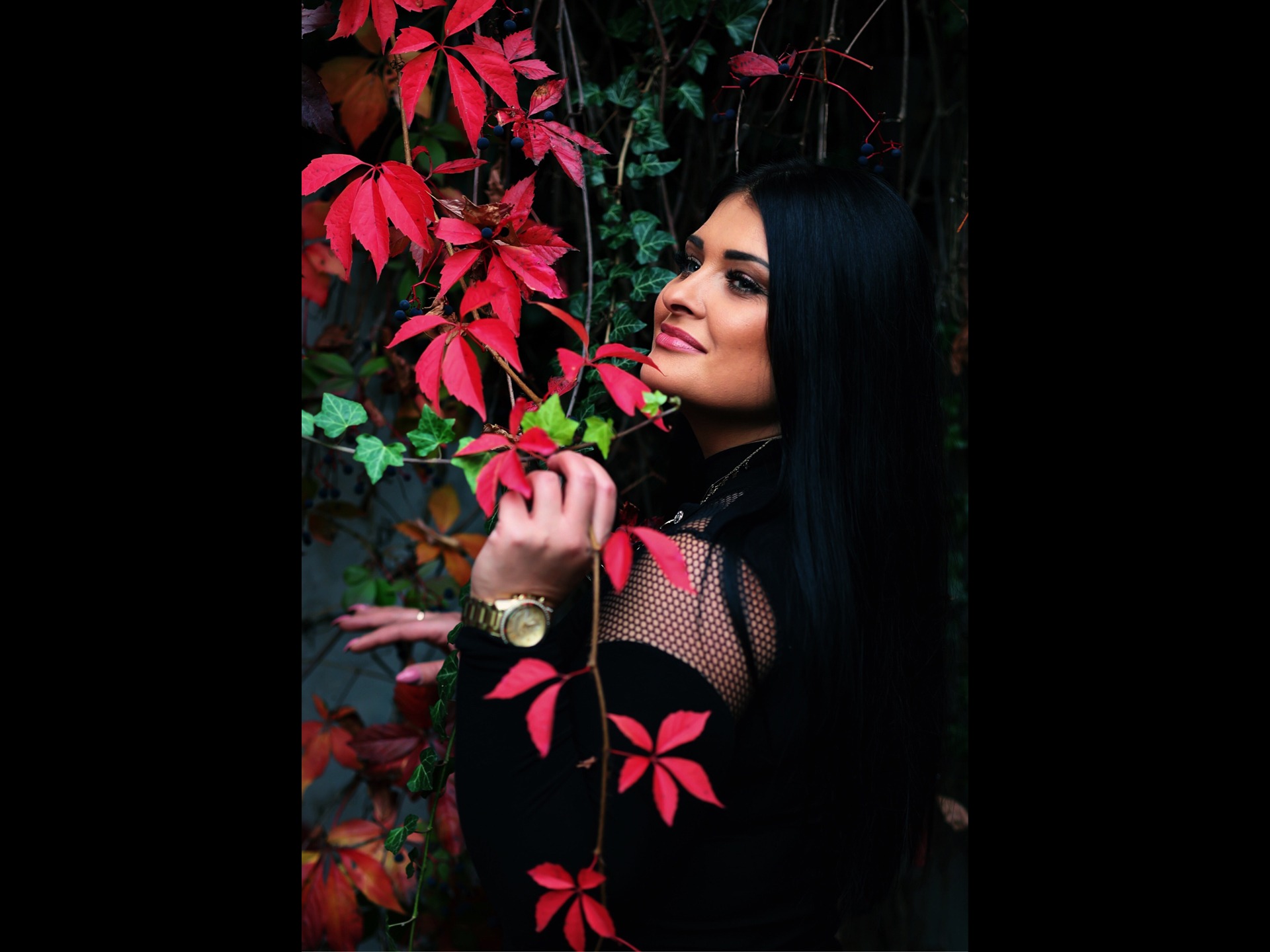 Portret uśmiechniętej kobiety z długimi czarnymi włosami, pozującej przy ścianie z czerwonych i zielonych liści, widoczny złoty zegarek na ręce.