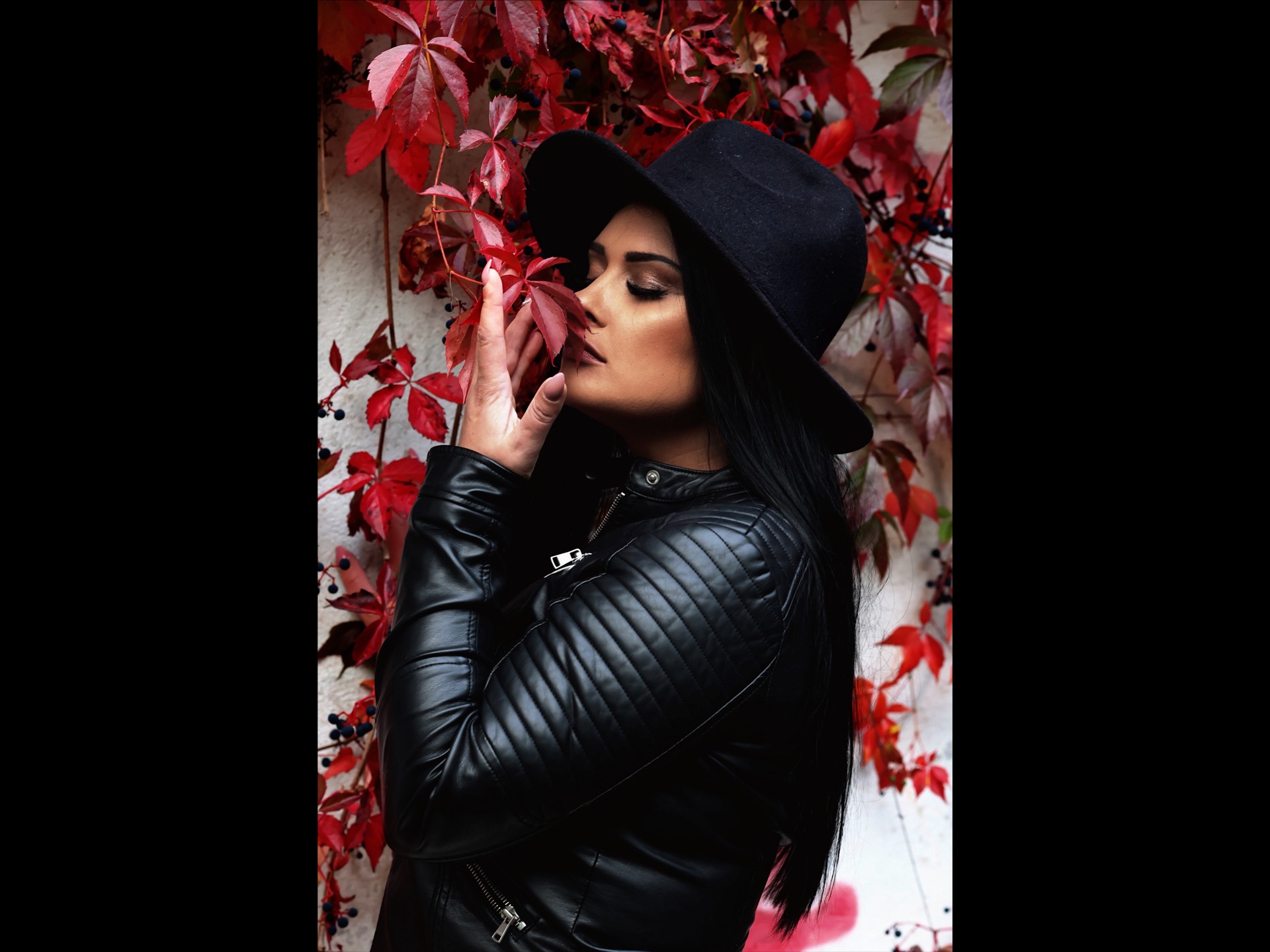Kobieta w czarnym kapeluszu i skórzanej kurtce pozuje z liściem winobluszczu na tle ściany pokrytej winoroślą.