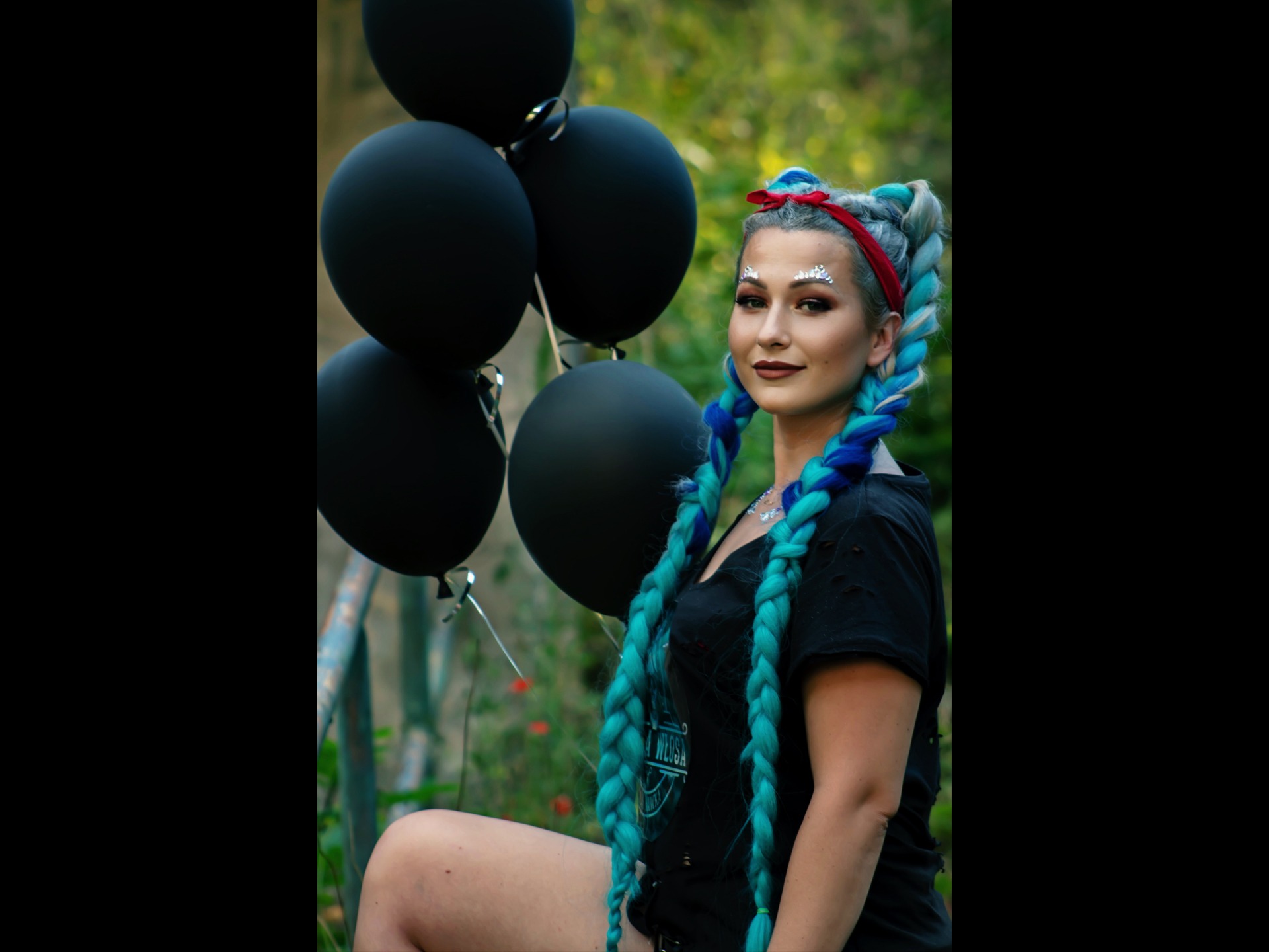 Portret kobiety z turkusowymi warkoczami, srebrnymi naklejkami na czole, trzymającej czarne balony na tle zieleni.