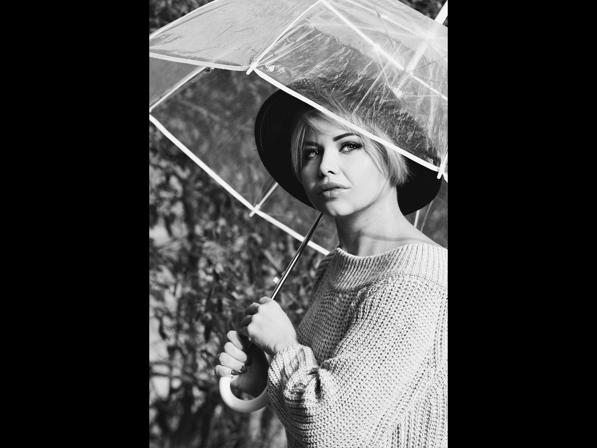 Portret kobiety w kapeluszu, trzymającej przezroczysty parasol na tle zarośli, zdjęcie czarno-białe.